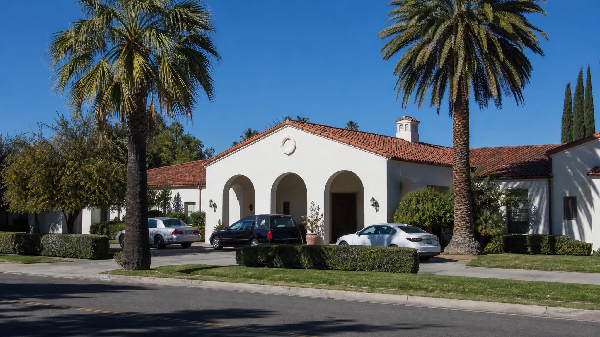 A single-story mission-style California funeral establishment in bright midday light โ white stucco walls, a red clay tile roof, a three-arched entrance portico, mature fan palms either side and a hearse waiting in the forecourt