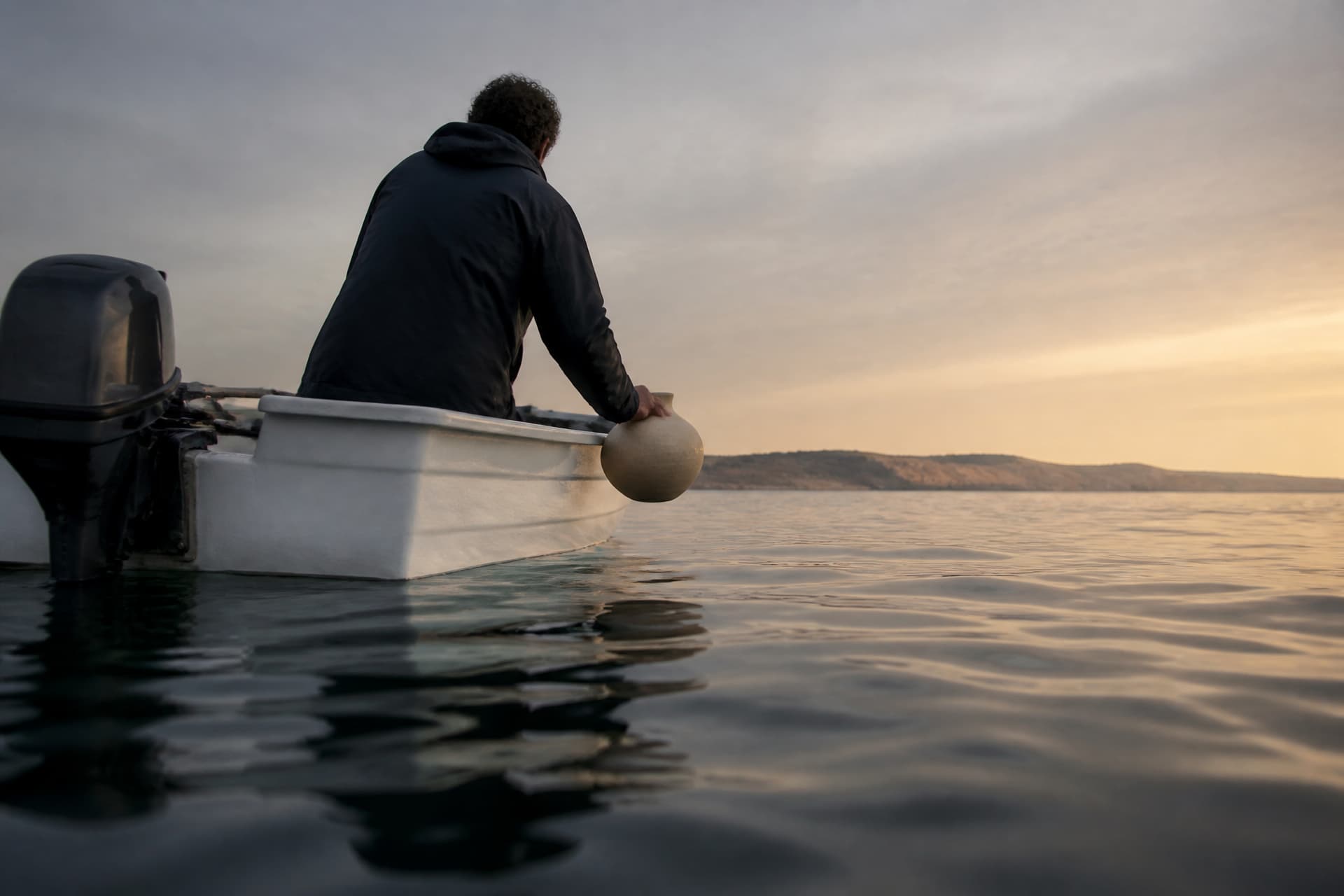 A small open outboard boat well out on the Pacific at first light, seen from water level, with one person seated at the stern in a dark jacket, seen from behind, holding a pale unglazed round vessel out over the side just above the water, and a low California headland far away on the horizon behind them