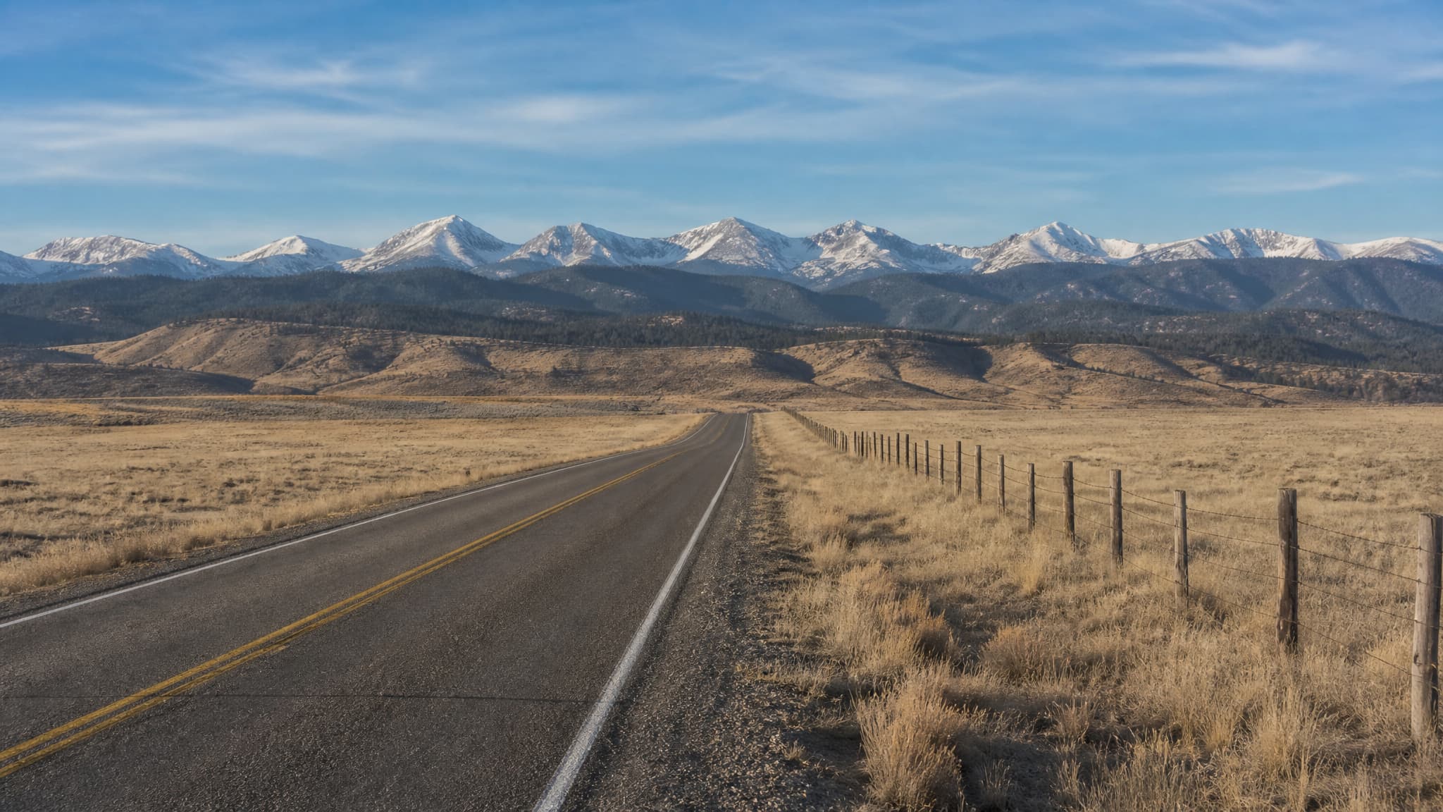 An empty two-lane road runs straight across tawny Colorado grassland toward the Front Range in clear morning light, a barbed wire fence along its right shoulder, tan foothills behind and snow still on the high peaks