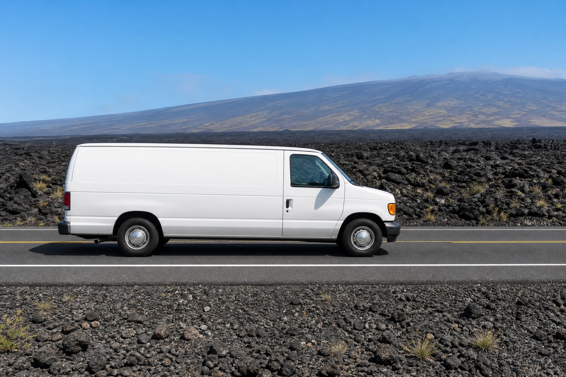 A two-lane highway crossing a black lava field on Hawai'i Island under bright sun, a plain white unmarked van in side profile in the right-hand lane, with the long shallow slope of a volcano on the horizon behind