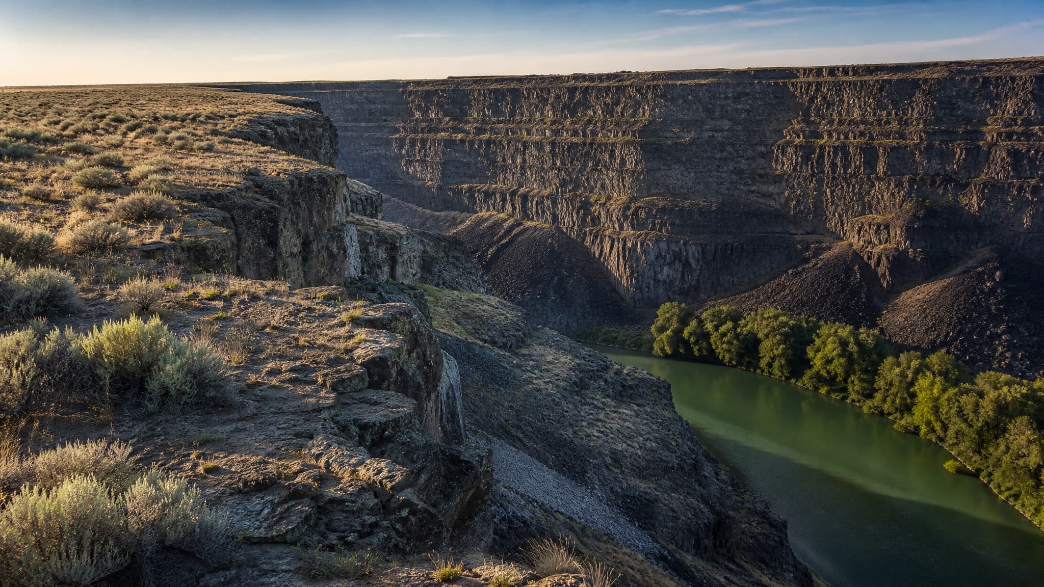 The rim of the Snake River canyon in low afternoon light, sheer stepped basalt walls dropping to a green river far below, flat sagebrush plain running unbroken to the canyon edge with no road, fence or building in frame