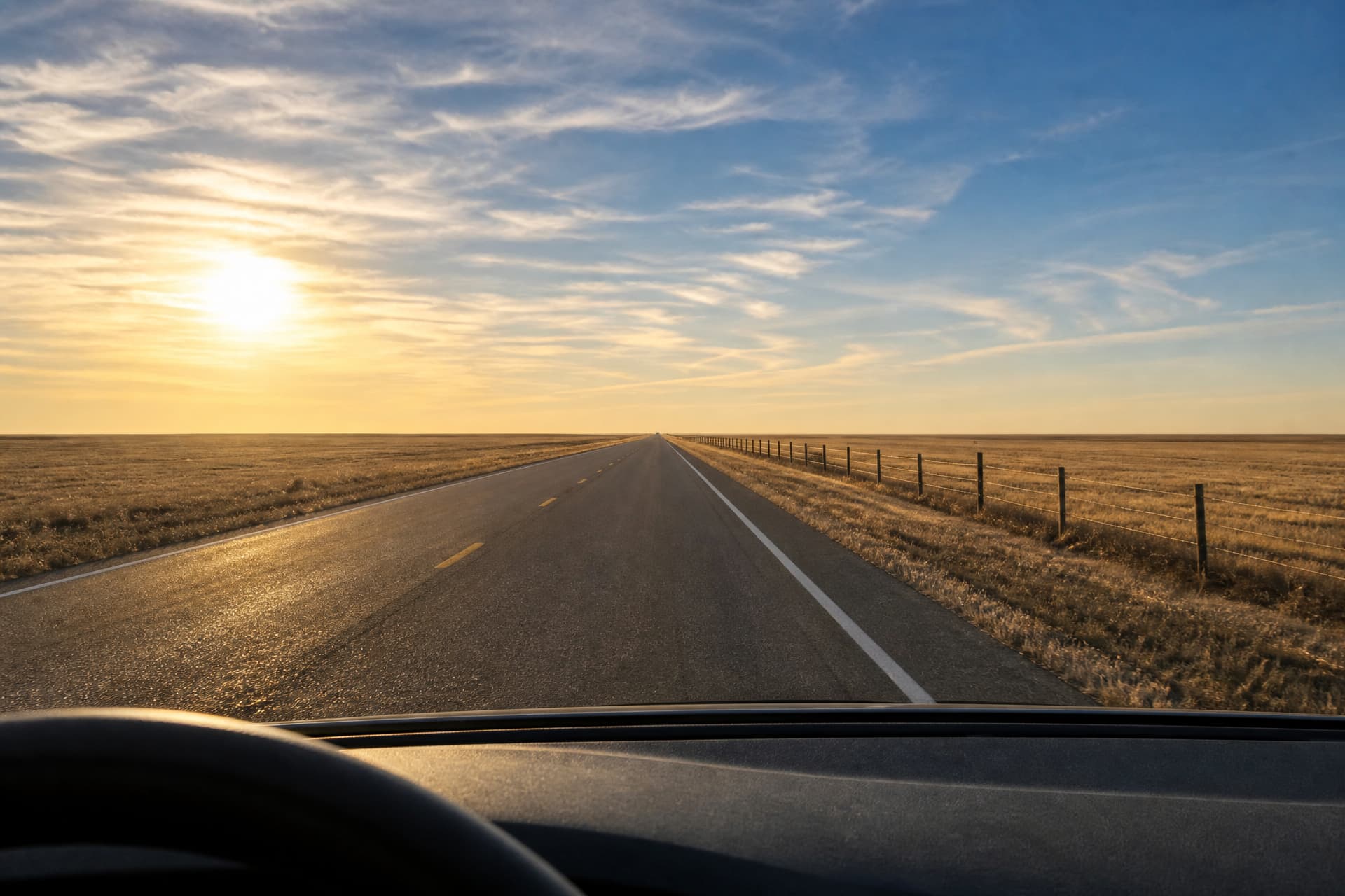 Seen from the driver's seat on a two-lane Kansas highway under a low sun, the wheel rim and dark dashboard across the bottom, the yellow center line to the left, a wire fence and gold stubble to the right