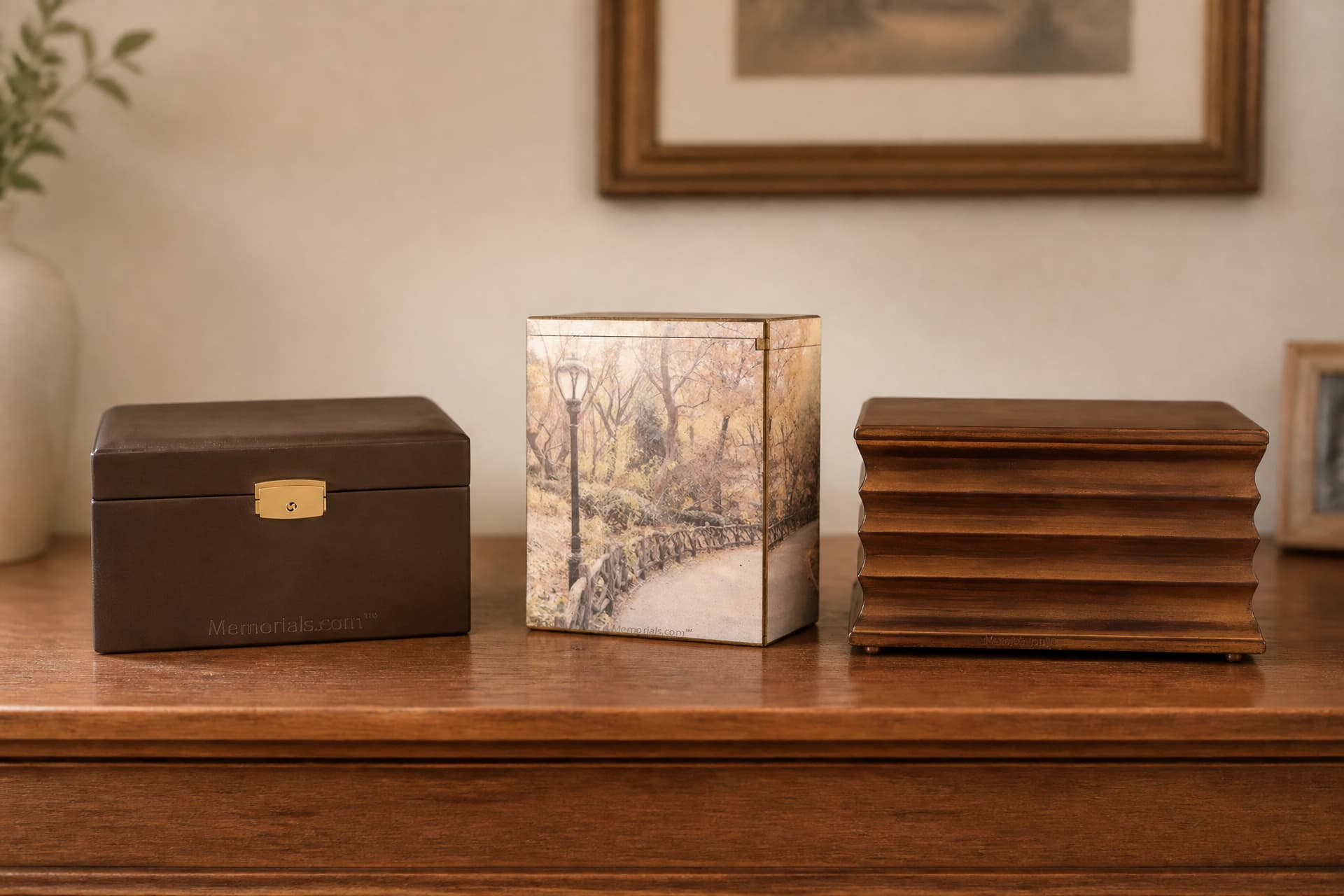 Three distinctive cremation urns spaced along a wooden sideboard in soft daylight โ a brown leather box with a gold clasp, a tall urn wrapped in a woodland-path photograph, and a horizontally fluted wooden urn