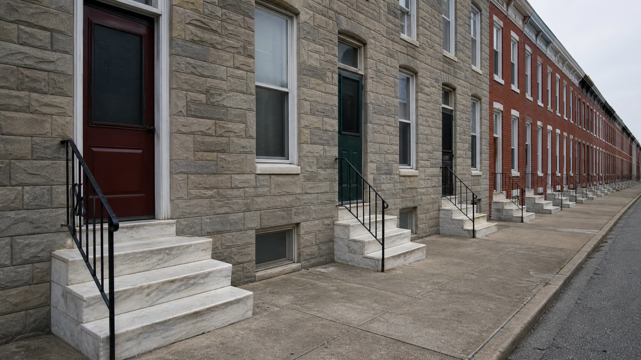 A Baltimore block of two-story rowhouses in flat overcast light, the near houses faced in gray formstone and the rest in red brick, white marble stoops stepping down the sidewalk one after another