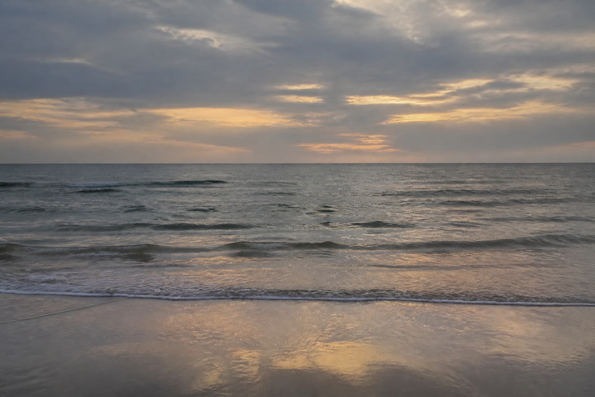 An empty Lake Michigan shoreline at dusk photographed from ground level, low waves running up onto wet sand that reflects the gray and gold sky, and a wide open horizon with no far shore, no boats and no people