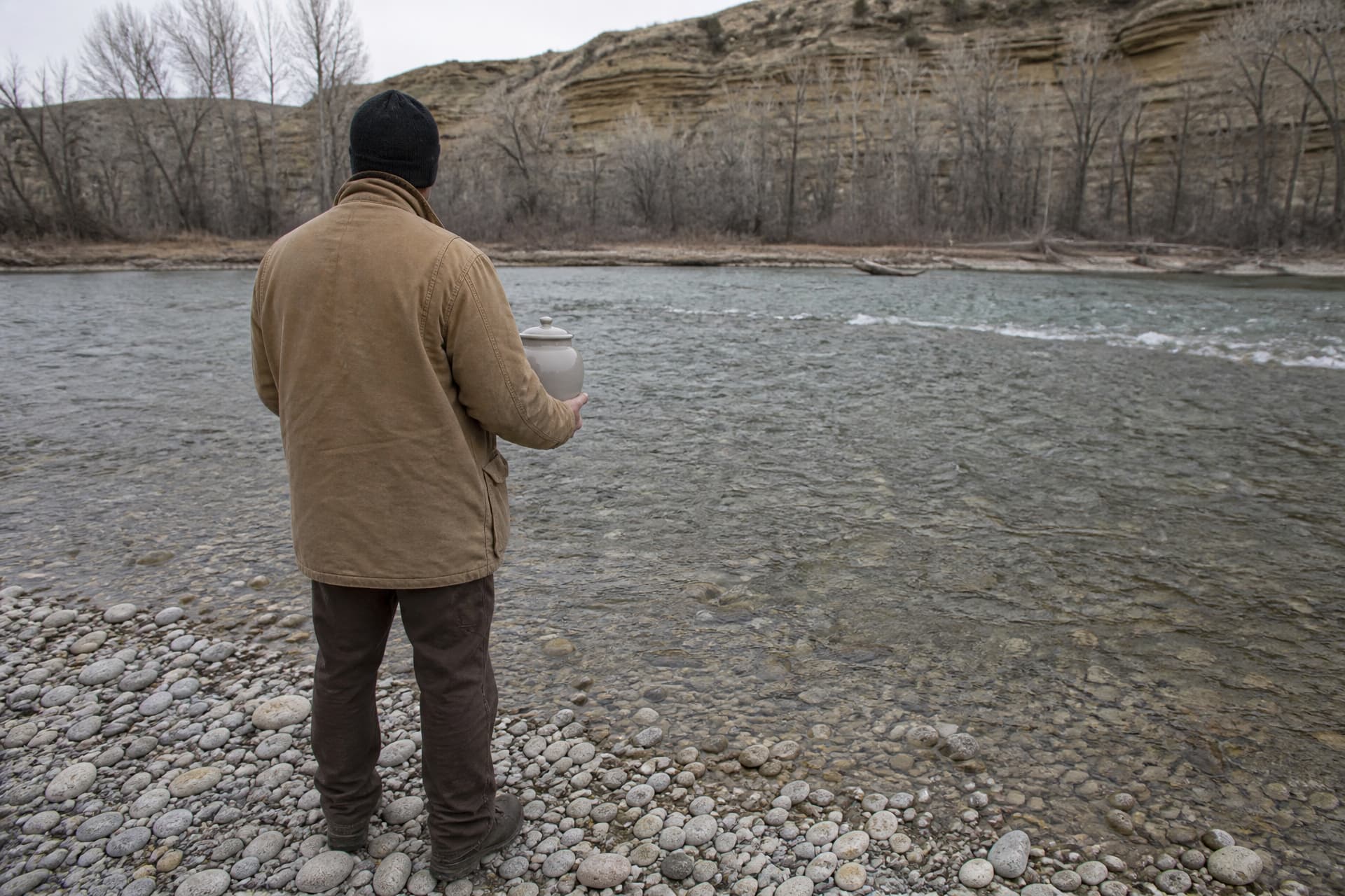 A person in a tan canvas coat, back to the camera on a cobble bar at the edge of a wide Montana river, cradling a plain pale ceramic lidded urn, bare cottonwoods and a banded sandstone bluff behind