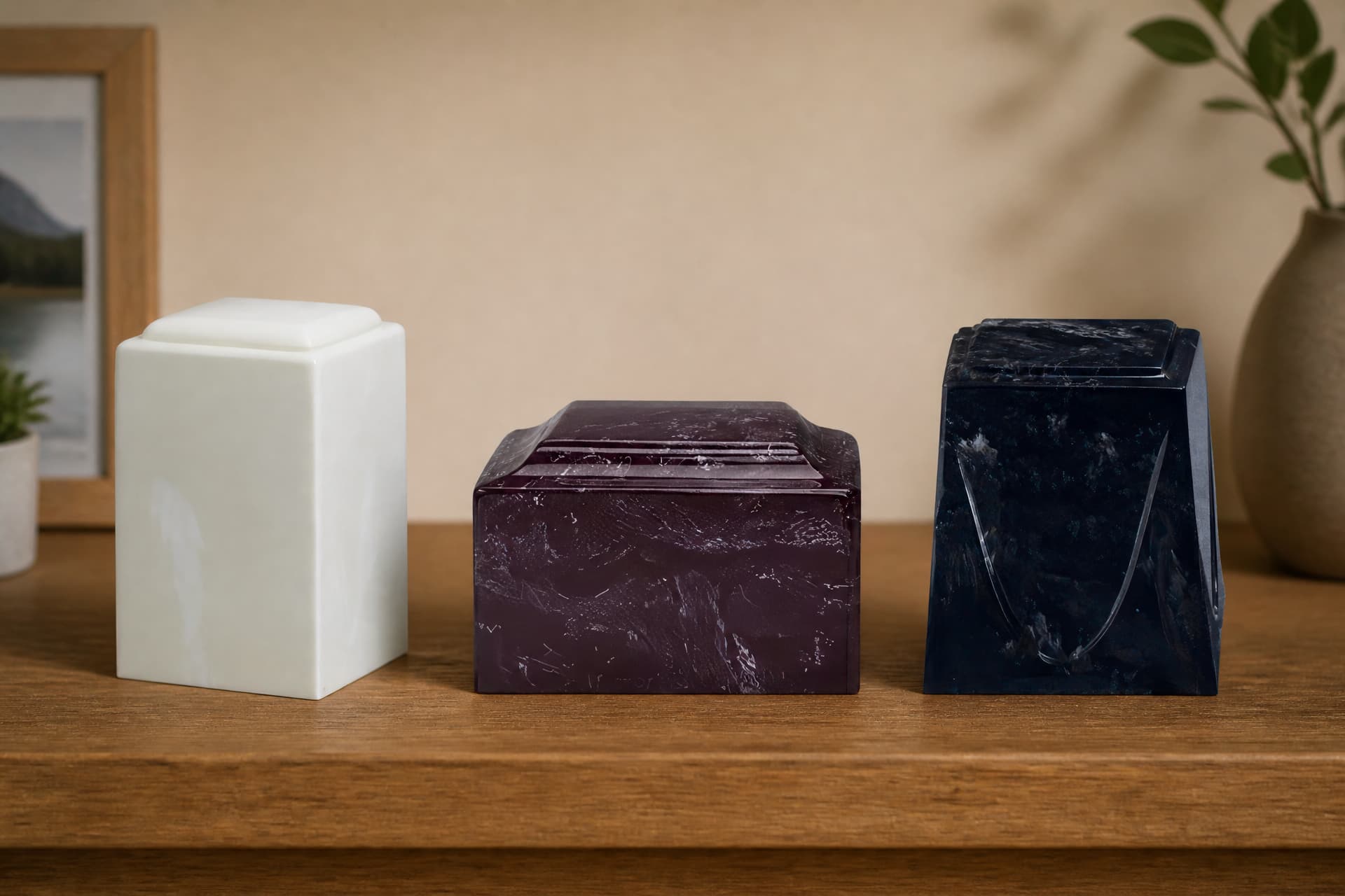 Three cultured stone cremation urns standing in a row on a wooden sideboard in warm indoor light: a plain cream square, a deep burgundy box with a stepped lid, and a near-black navy faceted urn