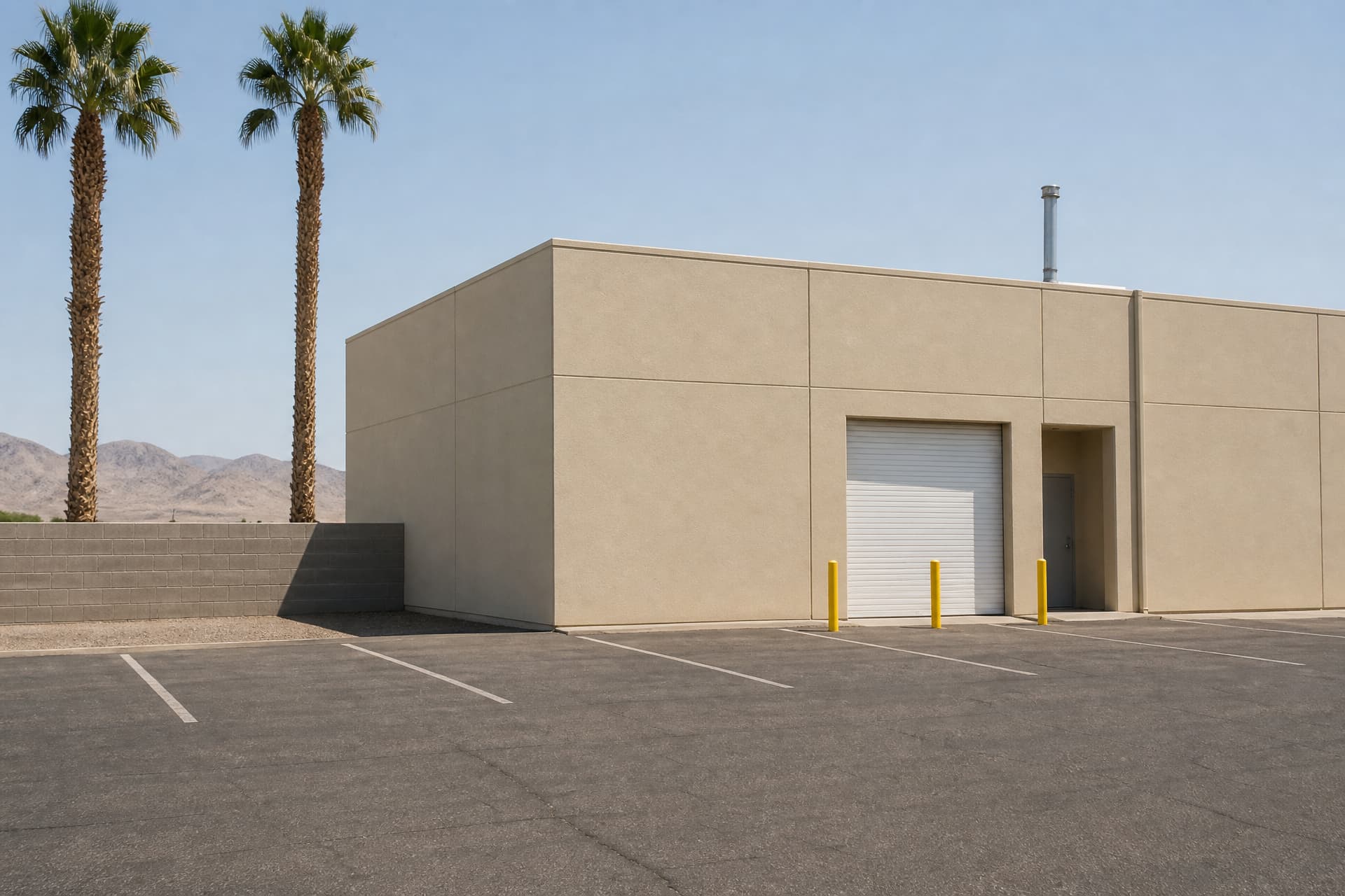 A beige concrete tilt-up building in a North Las Vegas industrial park under hard midday sun, a closed white roll-up bay door beside a shaded entry recess, yellow bollards on empty striped asphalt, brown mountains behind