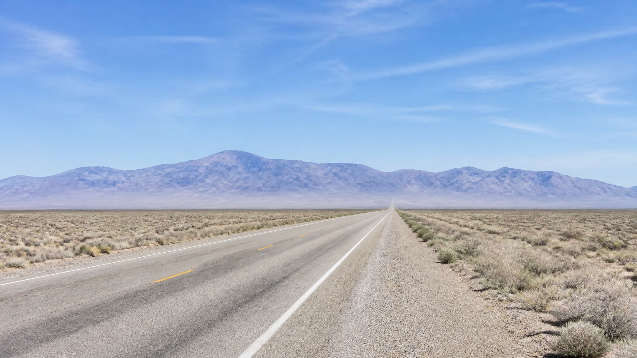 A wide Nevada basin at midday, an empty two-lane highway running dead ahead across pale sagebrush toward a long blue range, dashed yellow center line converging, heat shimmer flattening the road at the horizon