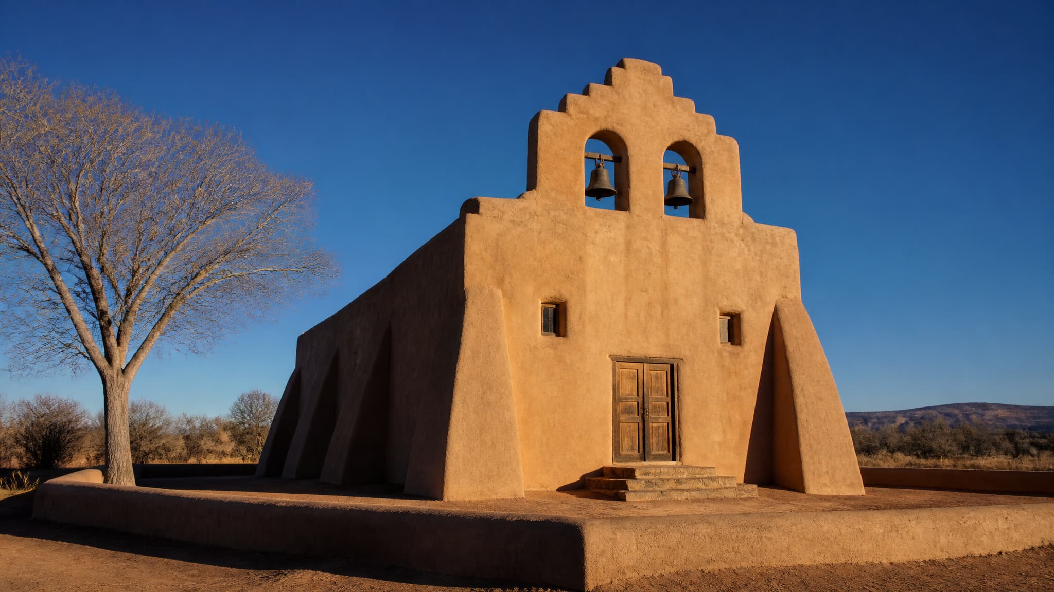 A New Mexico adobe church in late afternoon, thick earth-plastered walls warm against a hard blue sky, two bells hanging in the gable bell wall, and a long shadow thrown across the packed dirt yard