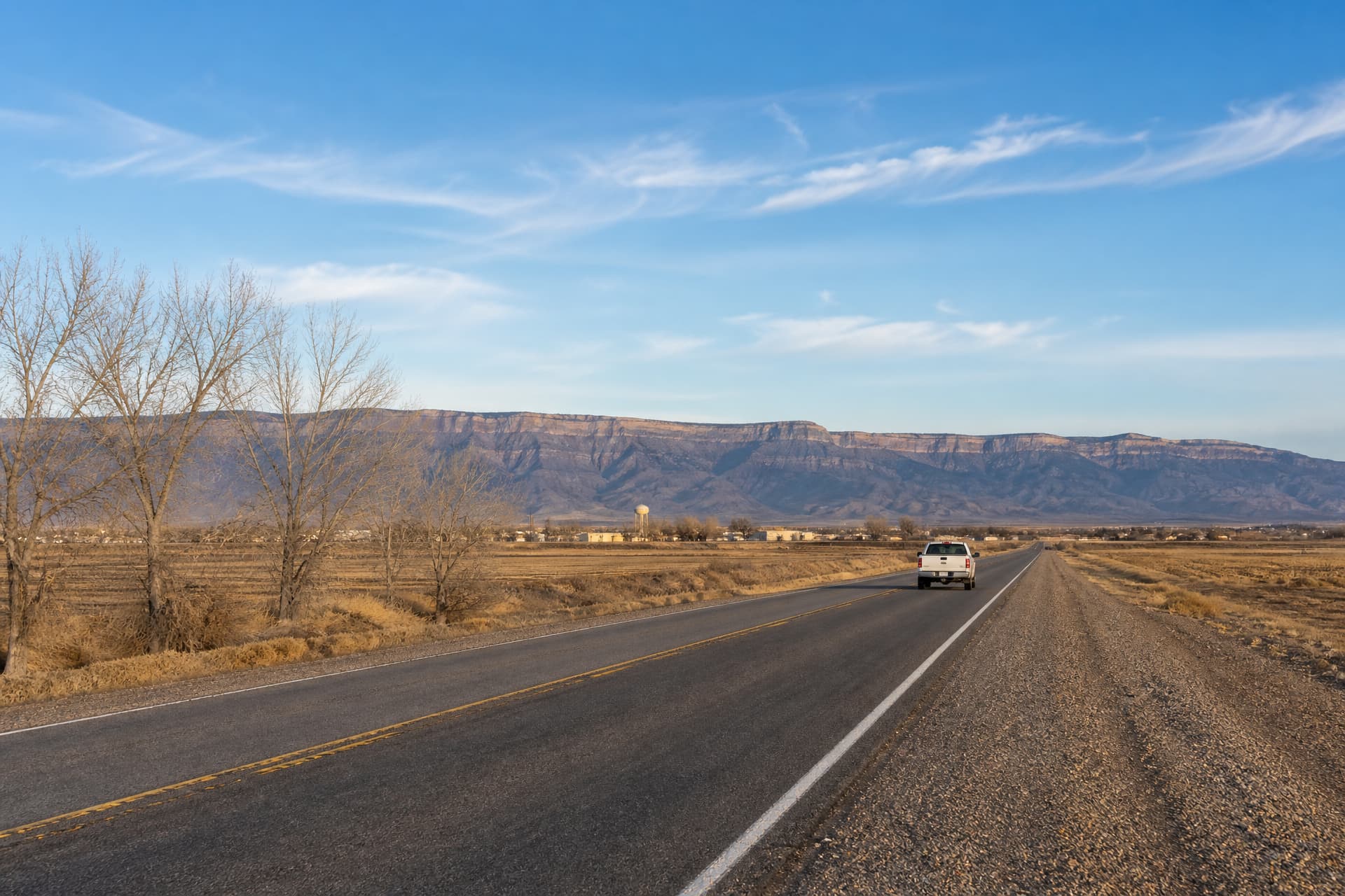 A two-lane New Mexico highway on the Rio Grande valley floor in late afternoon, one white pickup traveling away in the right-hand lane, bare cottonwoods and winter fields to the left, a small town and a long escarpment beyond