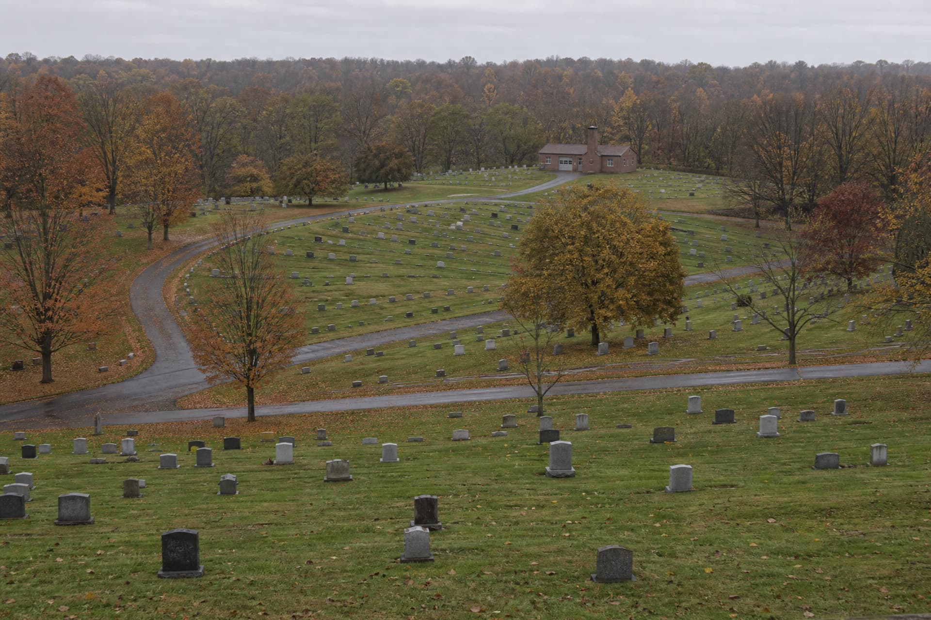 An upstate New York cemetery seen from a rise on an overcast autumn afternoon, wide and from above: mown burial sections divided by curving drives fill most of the frame, and a low crematory building with a square chimney stands small among them near the far treeline, a single access road running to it