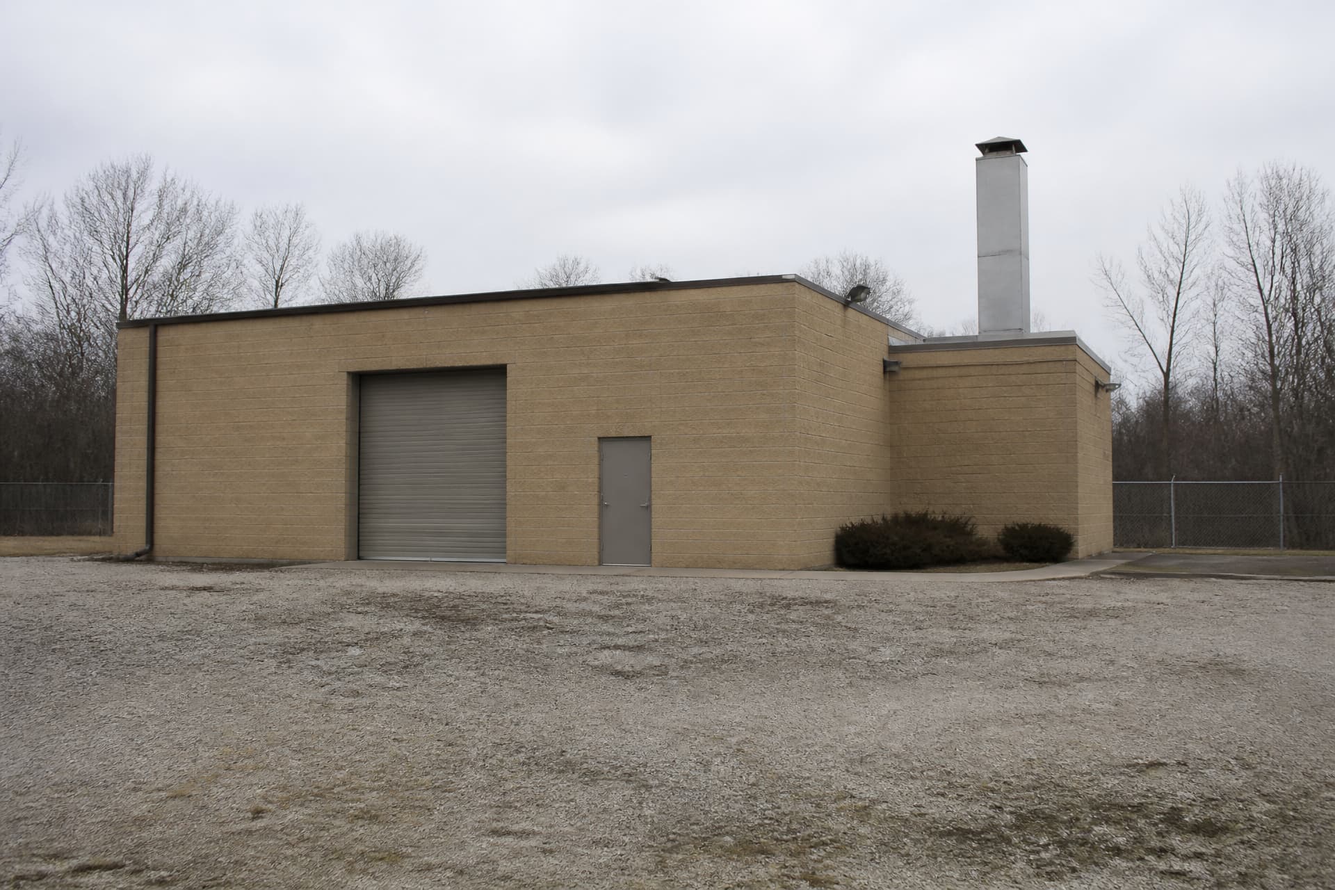 A plain single-story crematory building in buff-colored brick on a gravel service yard, with a roll-up vehicle door, one steel personnel door and a tall stainless steel exhaust stack rising from the roof, bare winter trees behind
