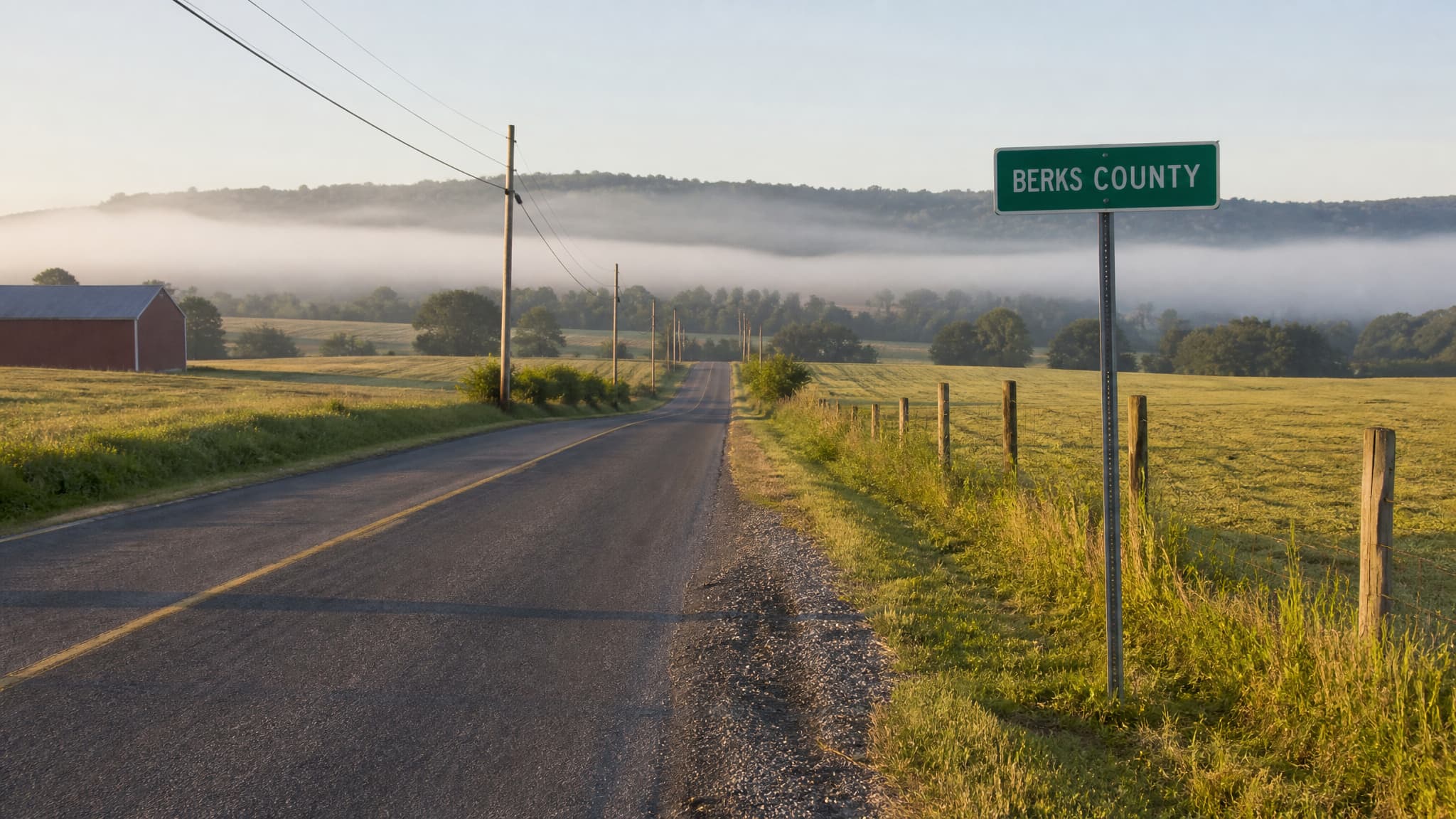 A two-lane Pennsylvania country road crossing a county boundary in early morning, with a plain green road sign reading BERKS COUNTY at the roadside, rolling farmland and a low mist behind it