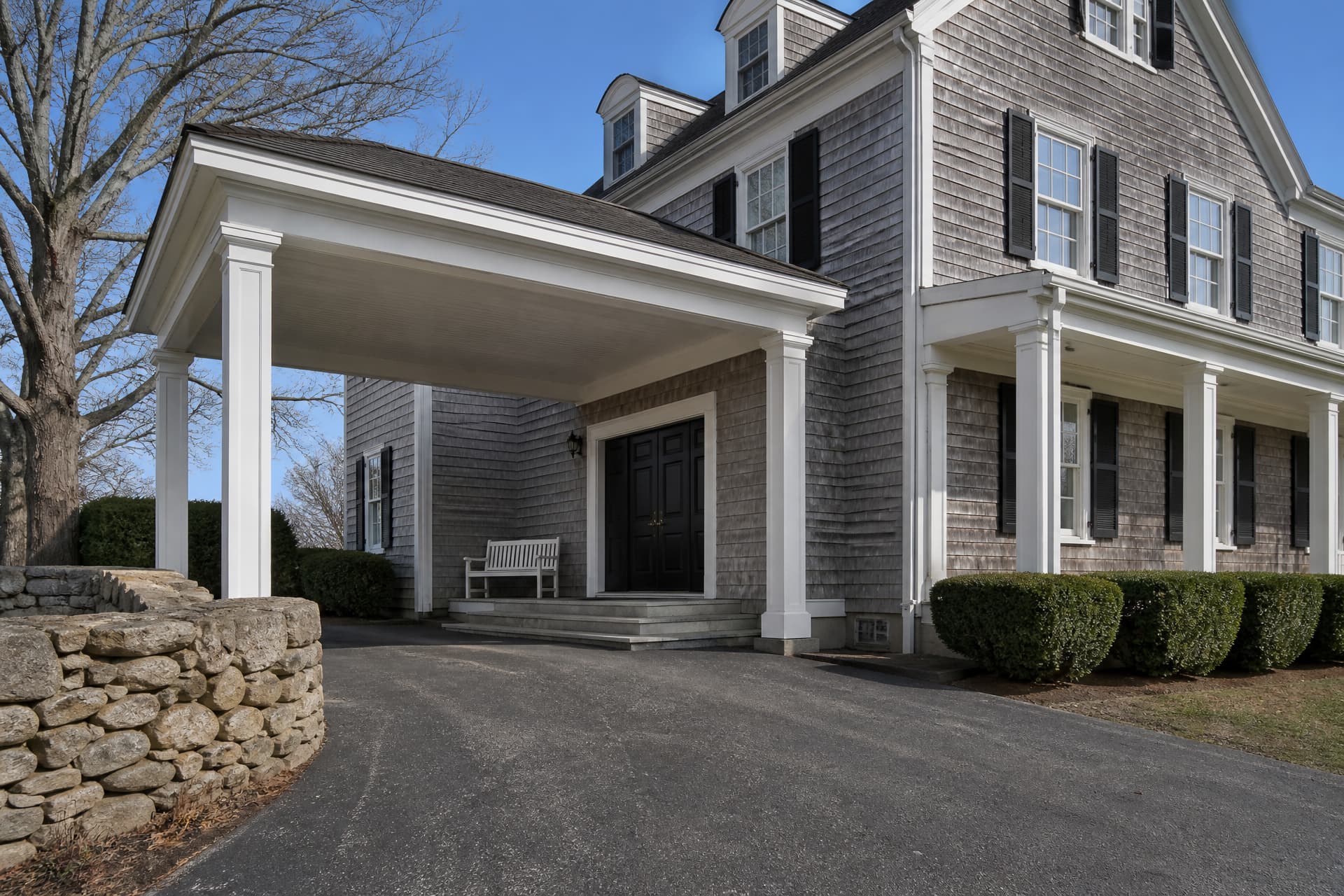 A gray shingled New England funeral home in morning sun, its white-columned porte-cochere over the driveway sheltering granite steps to a black paneled door and a white bench, with a fieldstone wall and a bare maple at the left