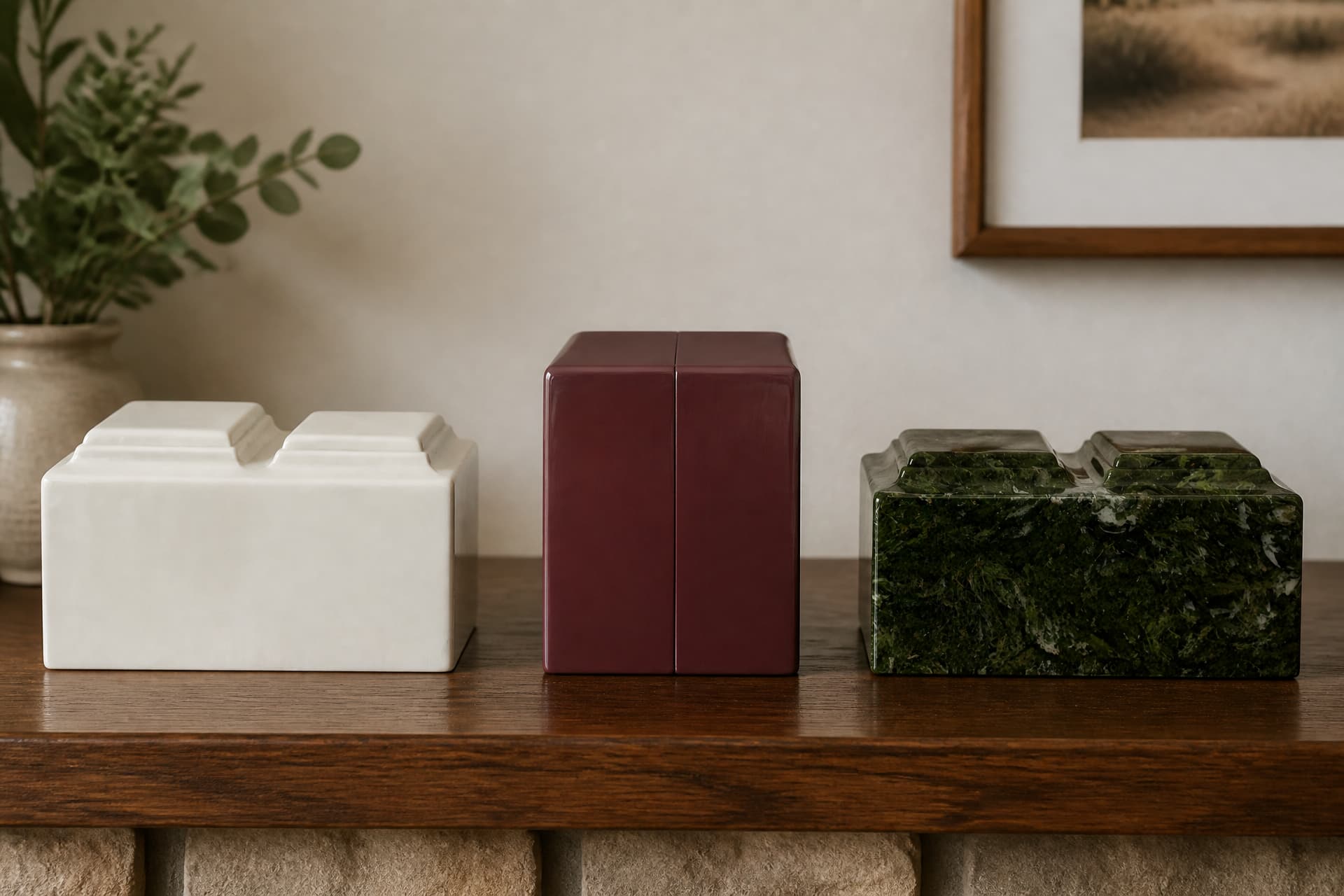 Three companion cremation urns in a row on a dark walnut sideboard: a cream twin-lid marble box, a glossy burgundy cube and a green marble twin-lid box, with a plant and a framed picture blurred behind
