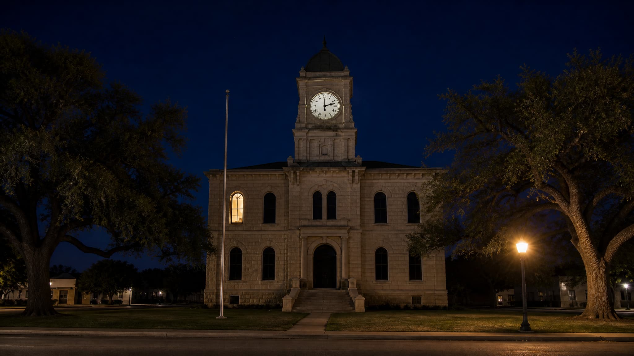 A small Texas county courthouse at night: a two-story cut-limestone building with a domed clock tower whose white Roman-numeral dial reads two o'clock, one upstairs window lit warm and every other window dark, a bare flagpole with no flag, live oaks on either side and a single lit street lamp, and no lettering or signage anywhere on it