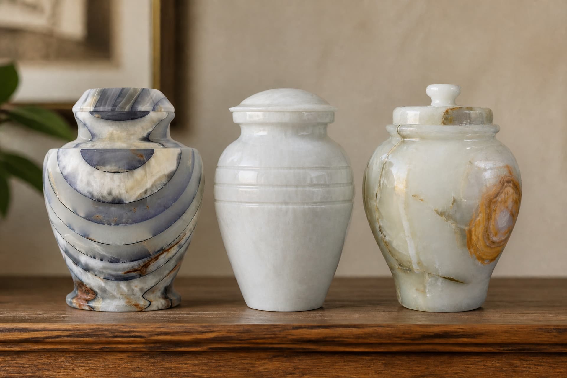 Three stone cremation urns in a row on a wooden shelf in soft daylight โ a banded blue-gray and cream urn, a plain white one with turned rings, and a pale onyx one with an orange swirl