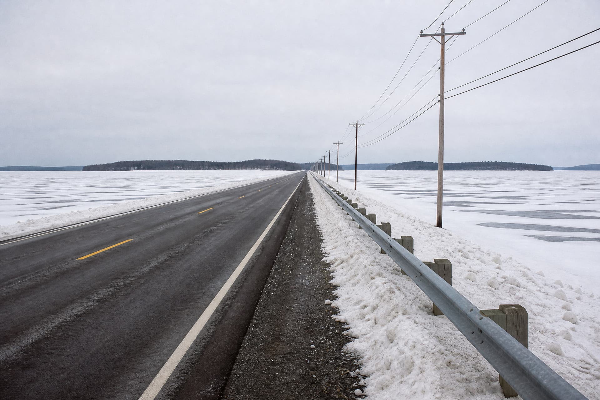 A two-lane causeway running straight out across a frozen lake toward low wooded islands in flat winter light, snow-covered ice on both sides, a guardrail and a line of utility poles receding beside it