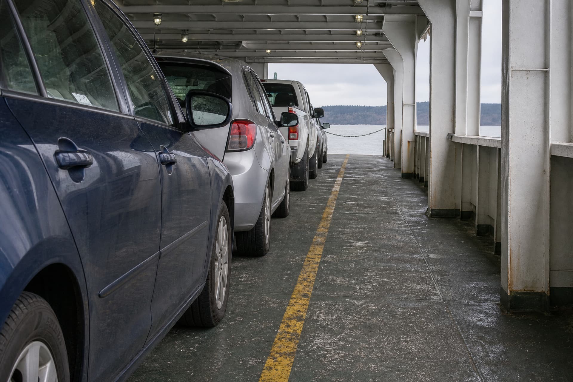 The car deck of a Washington State Ferry mid-crossing, seen from beside a row of parked cars at door height, looking forward past their flanks to the open end, gray water and a low forested shore