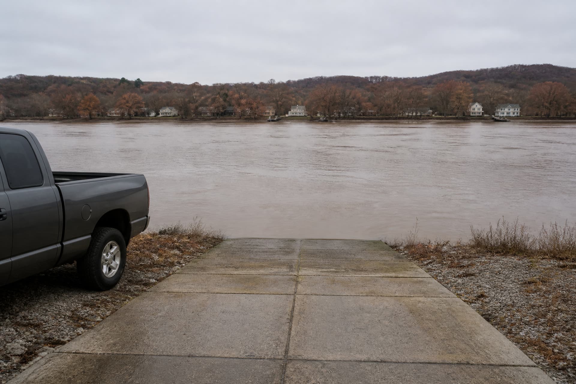 A concrete boat ramp running into a wide brown river under flat gray overcast, the back half of a parked gray pickup cut off at the left edge of the frame, and small pale houses along the far wooded shore