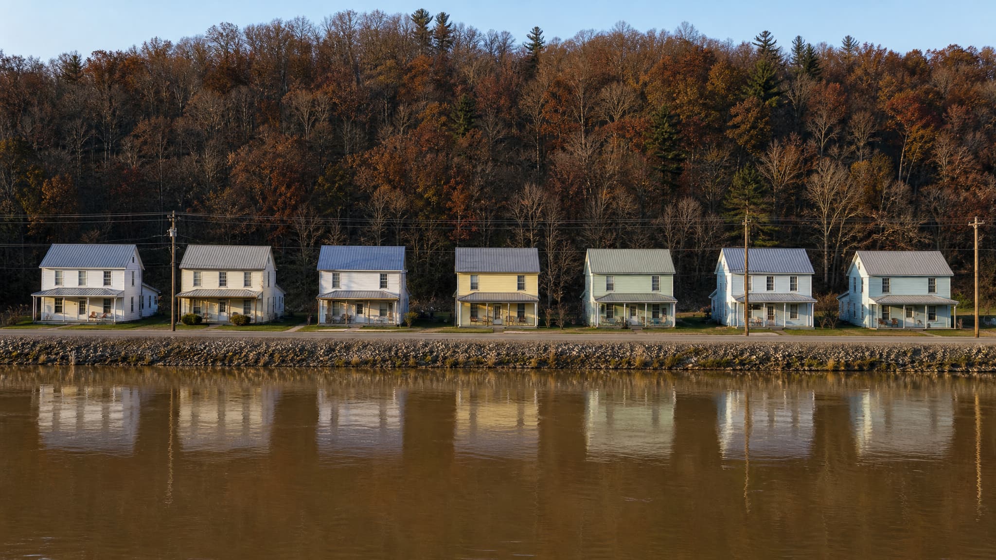 A row of seven plain two-story frame houses in white, cream and pale blue standing along a paved road on a narrow strip of ground between a wide brown river and a steep autumn ridge