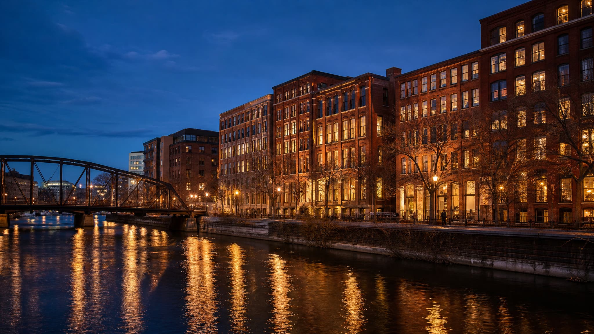 A row of restored red-brick warehouse buildings along the river in Milwaukee's Third Ward at dusk, their windows lit warm against a deep blue sky, with a steel bridge at the left and reflections breaking on the water