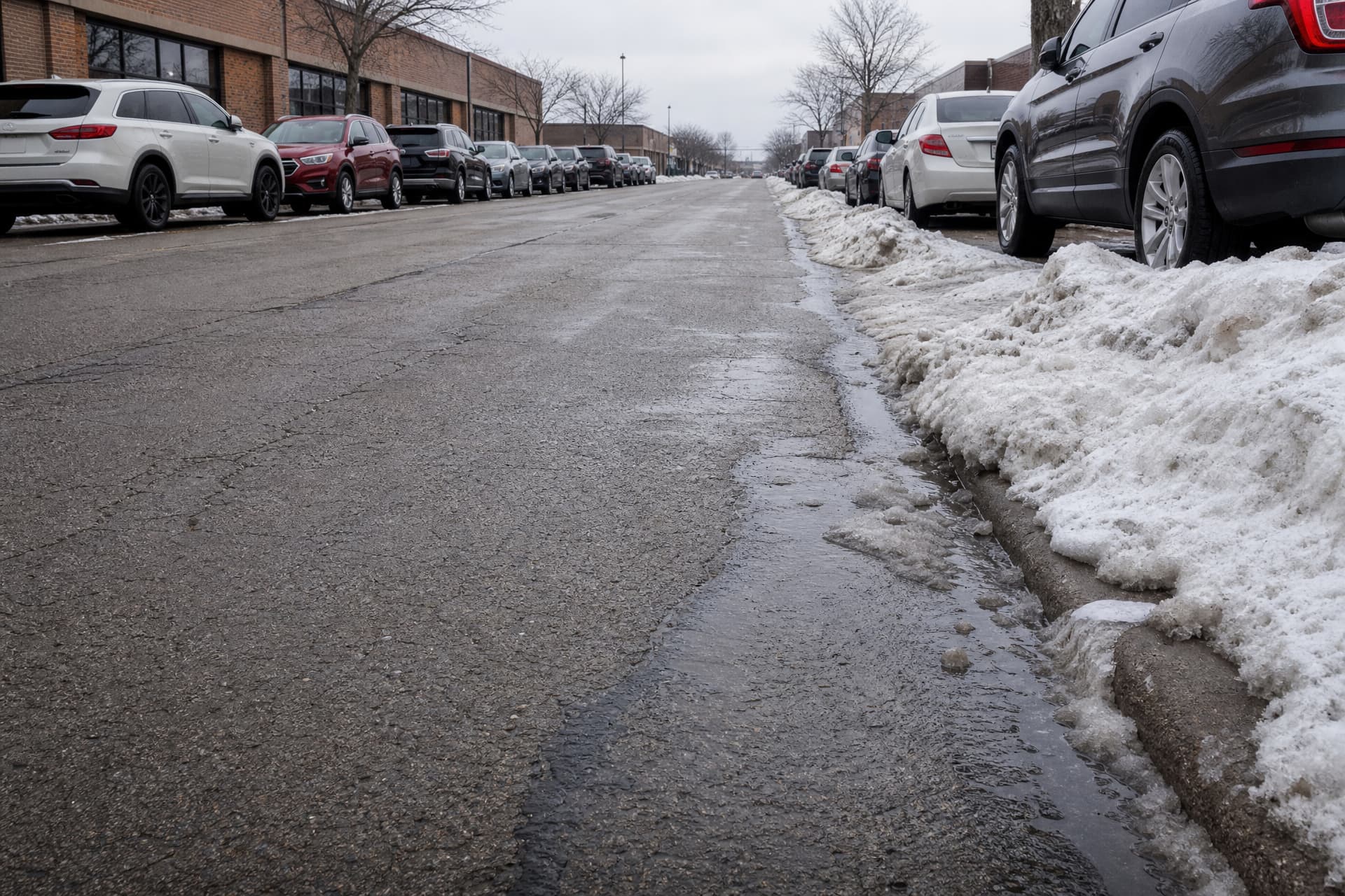 A wide suburban commercial street in West Allis in late winter, photographed low from the curb so the frame holds the road, the banked snow and the lines of parked cars, with the storefronts cropped below their sign bands