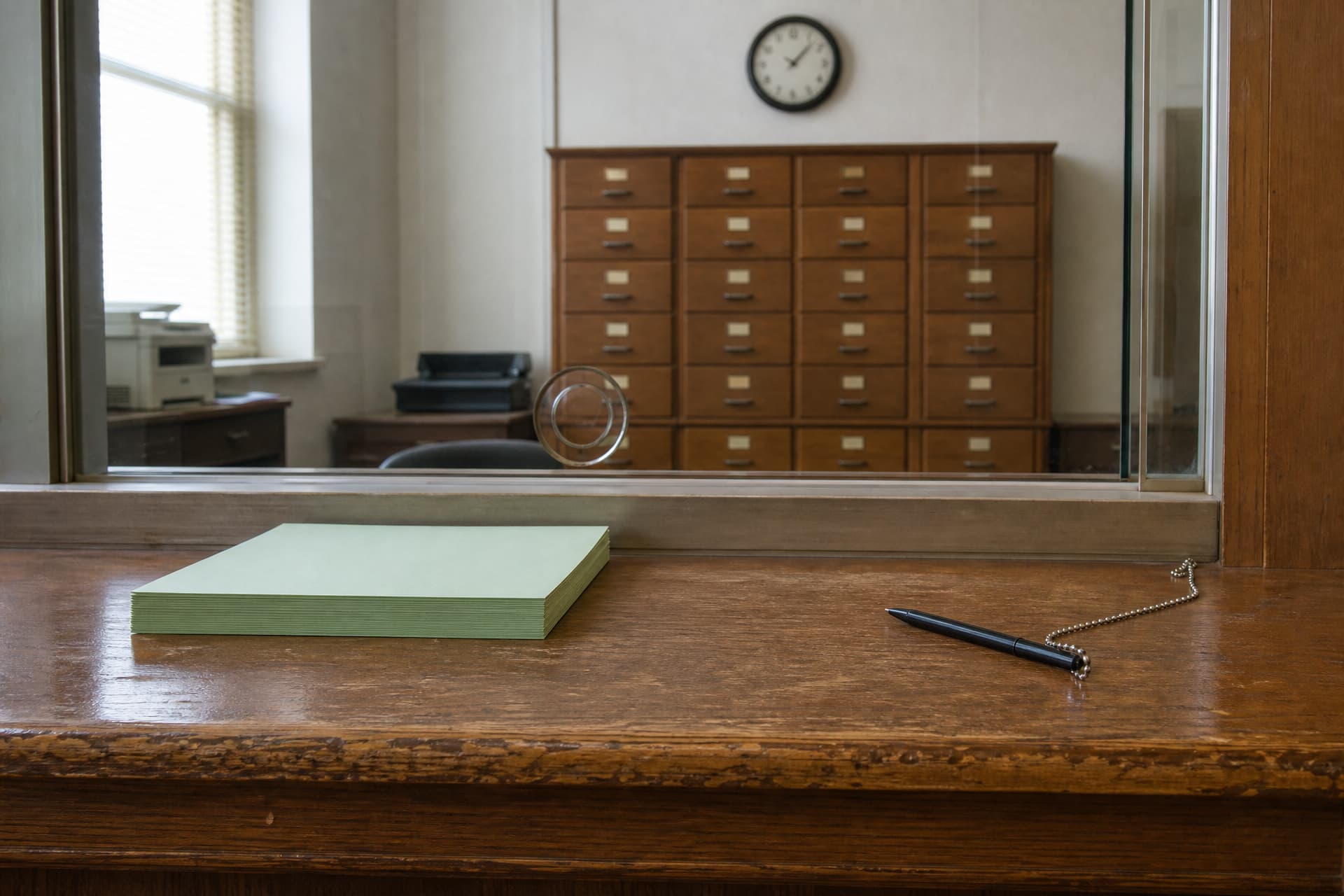 A Wisconsin county register of deeds counter with a stack of blank forms and a pen on a chain, a glass partition and filing cabinet behind