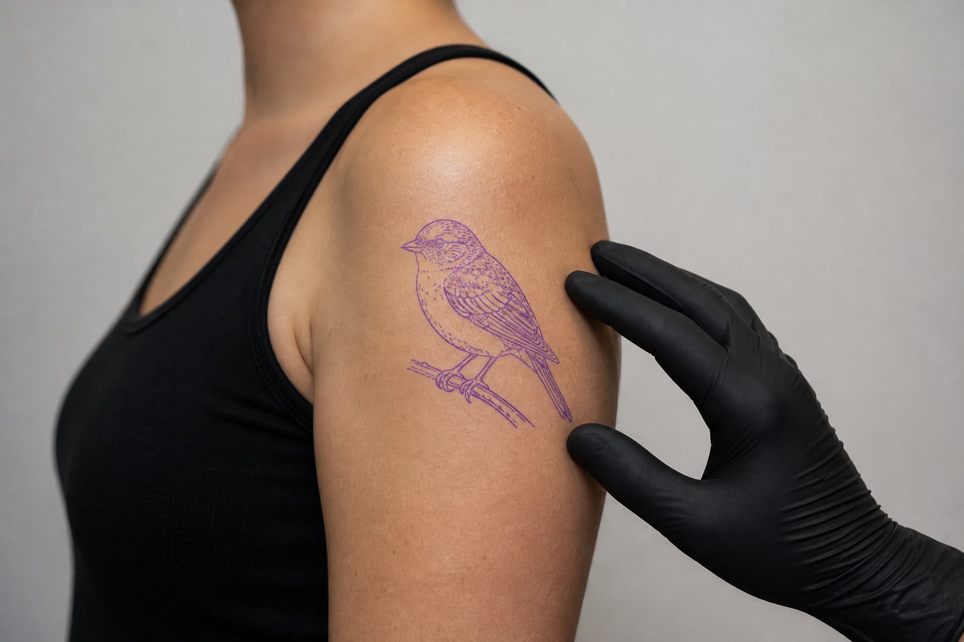 A tattoo stencil of a small bird positioned on a shoulder, seen from the side, with the artist's gloved hand adjusting the placement