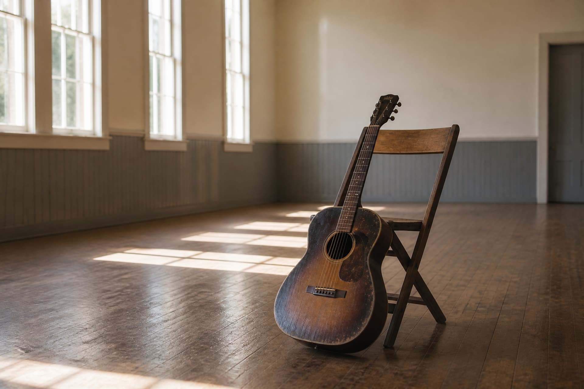 An acoustic guitar leaning against an empty wooden chair in a plain hall, late afternoon light falling across bare floorboards
