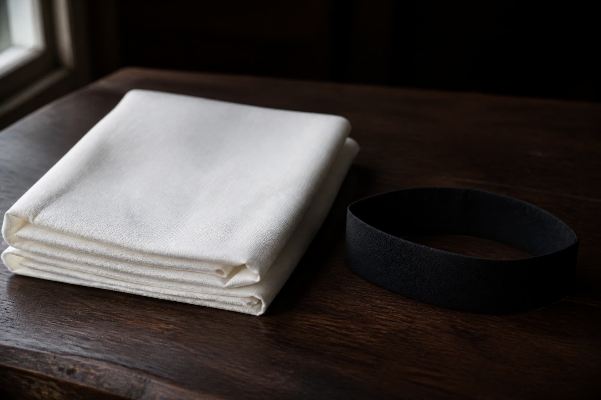 A folded length of undyed white cotton cloth on a dark wooden table in soft window light, with a black crepe mourning armband standing in a loop beside it