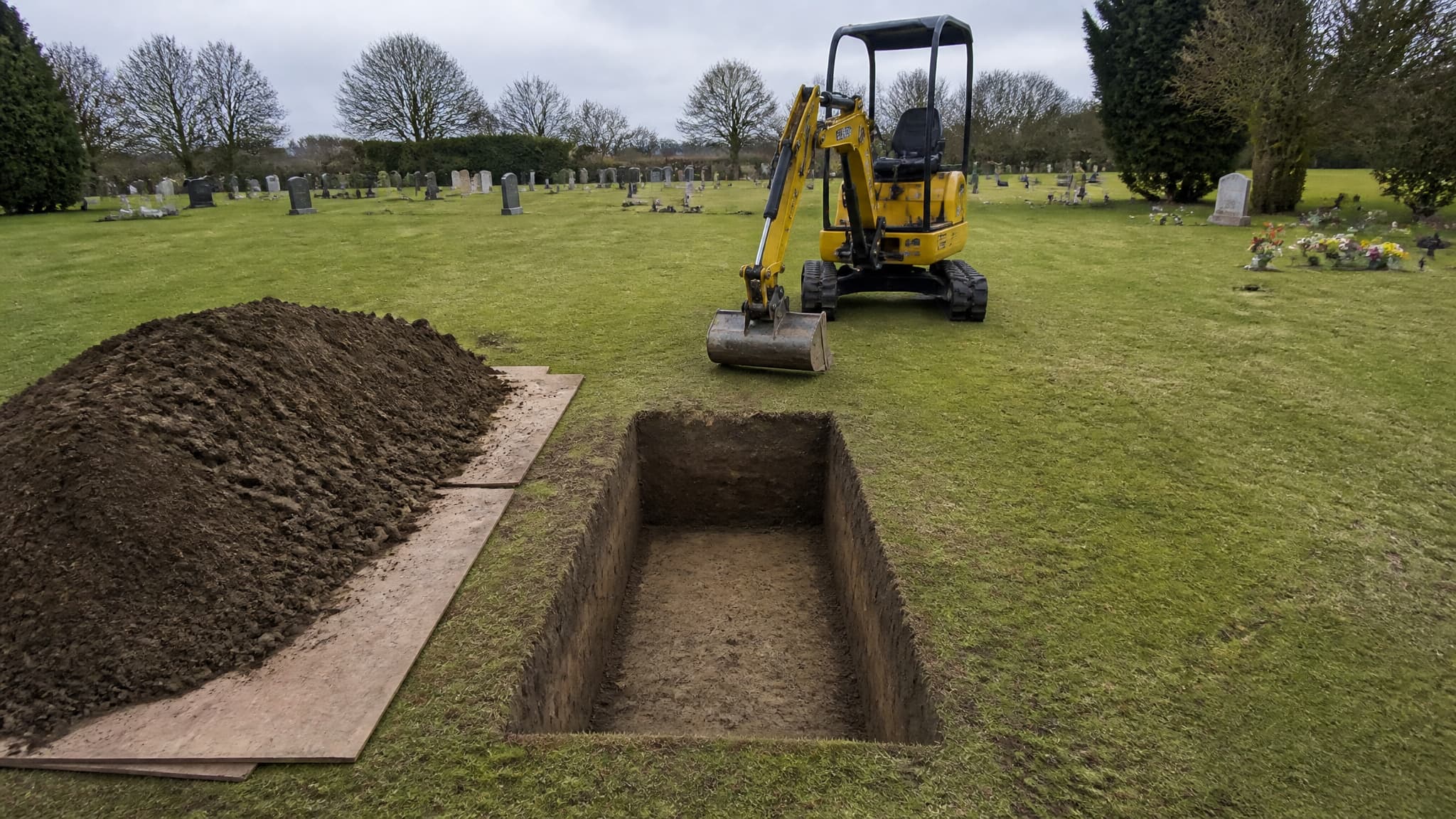 A freshly excavated grave in a cemetery with squared sides and a neat rectangular opening, the spoil heaped on plywood sheets to one side and a compact yellow excavator parked at the head of it in flat overcast light