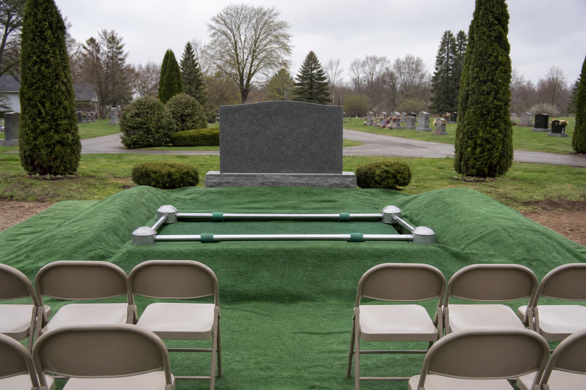 A grave dressed for a funeral service, the opening covered by a chrome lowering device with green artificial turf laid over the surrounding ground and the heaped soil, and two short rows of folding chairs facing it