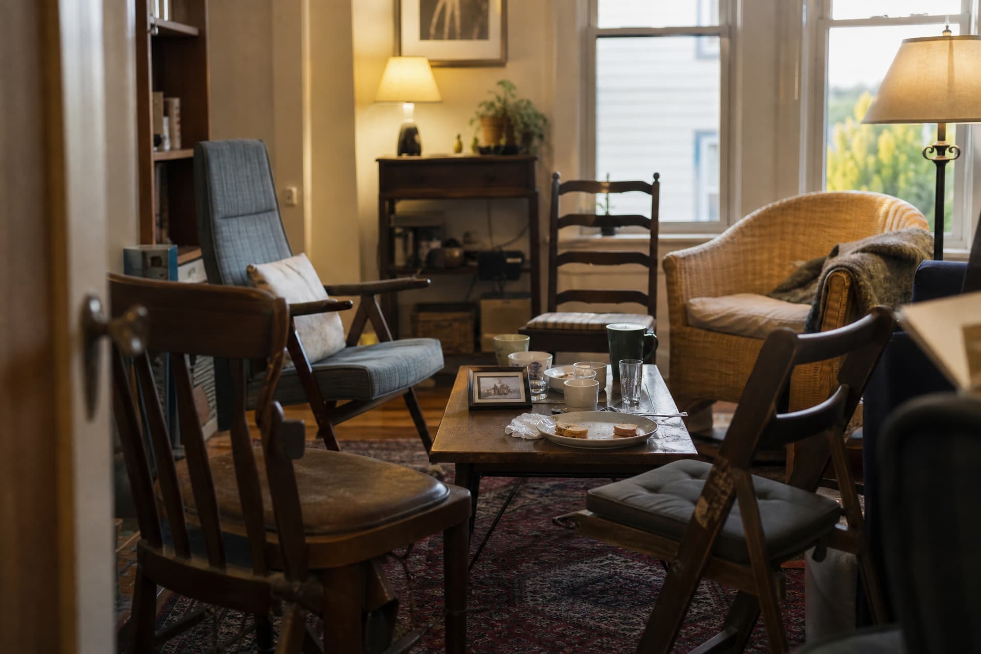 A domestic living room after a gathering, mismatched chairs pulled into a loose circle, used cups and a half-eaten plate of sandwiches on a low table and a framed photograph face up among them