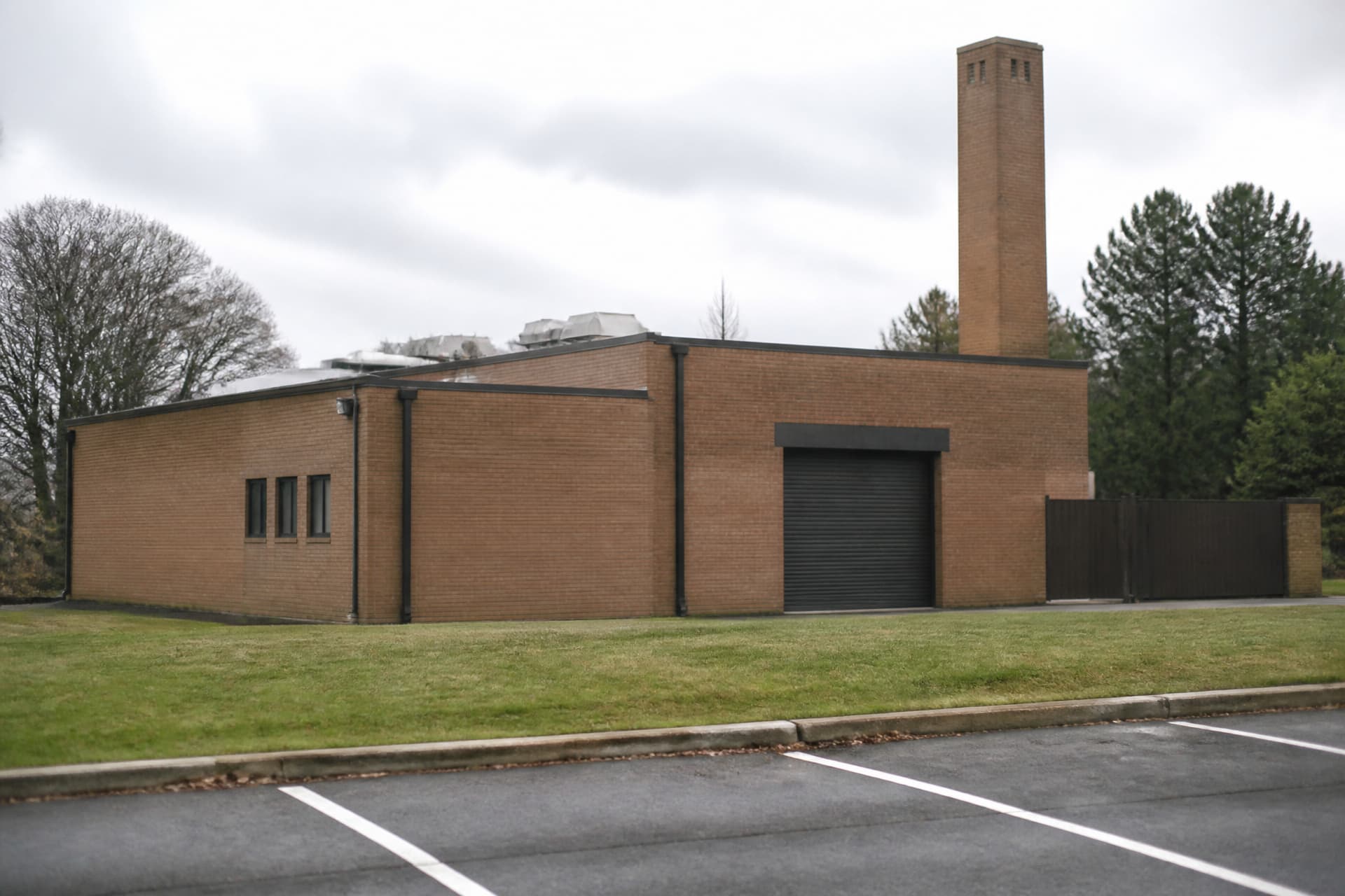 The plain exterior of a small crematory building on an overcast morning, low brick with a tall chimney and a single closed roller door, seen from an empty car park