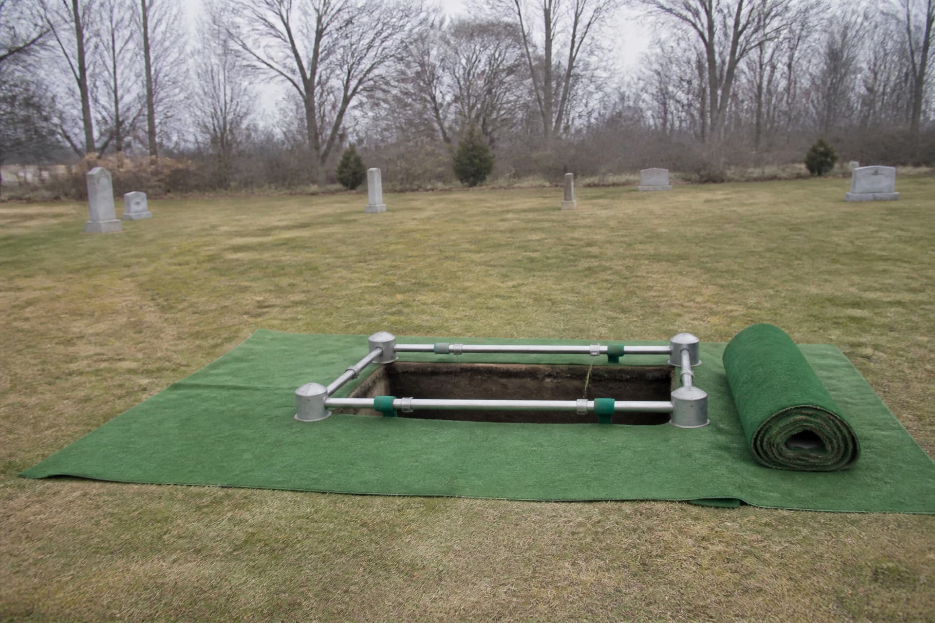 A prepared open grave with a plain lowering frame over it and a roll of artificial turf at one side, no chairs, no canopy and no flowers, mown cemetery grass and bare trees behind