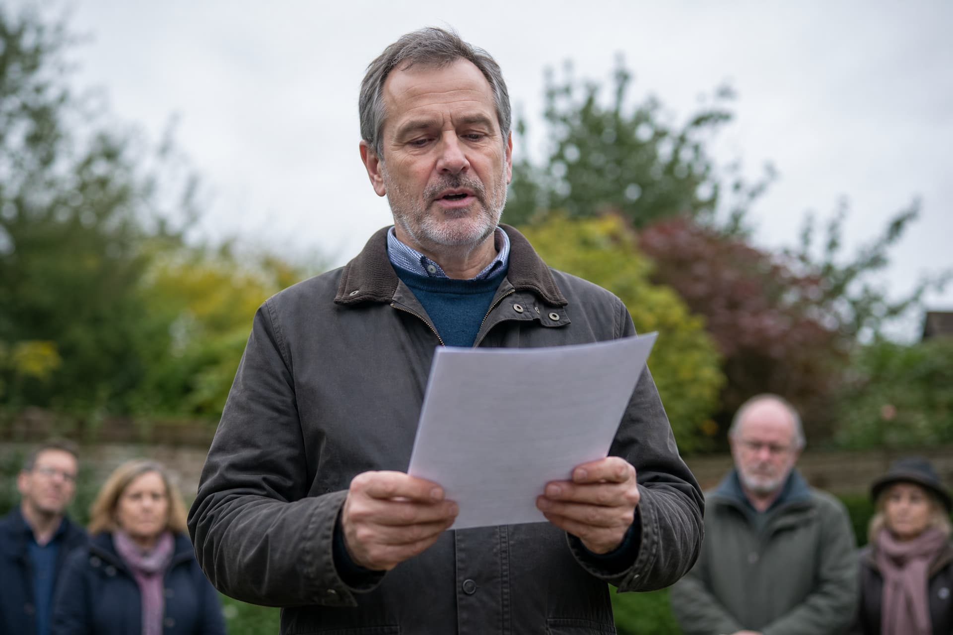 A man reading aloud from a printed sheet held in both hands, outdoors, listeners blurred behind him