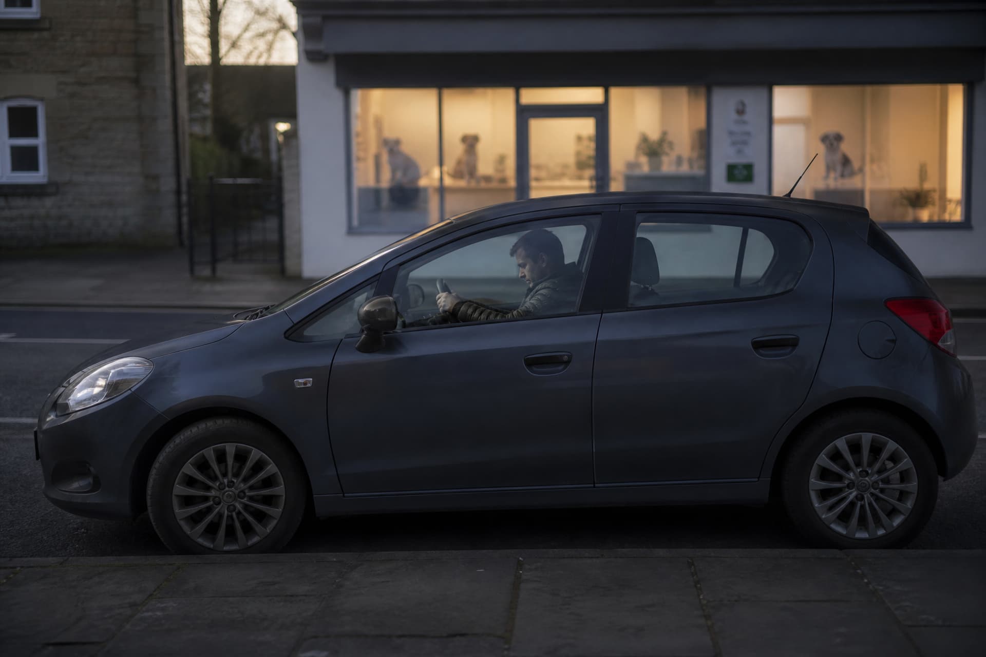 A person sitting in a parked car at dawn with both hands on the wheel, seen through the side window