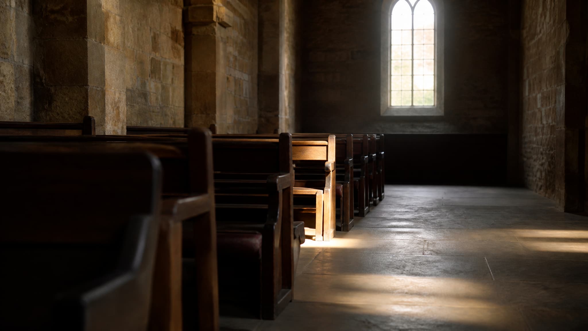 The back rows of an empty church in soft daylight, wooden pews receding toward a bright window, one pew near the aisle catching the light more than the others