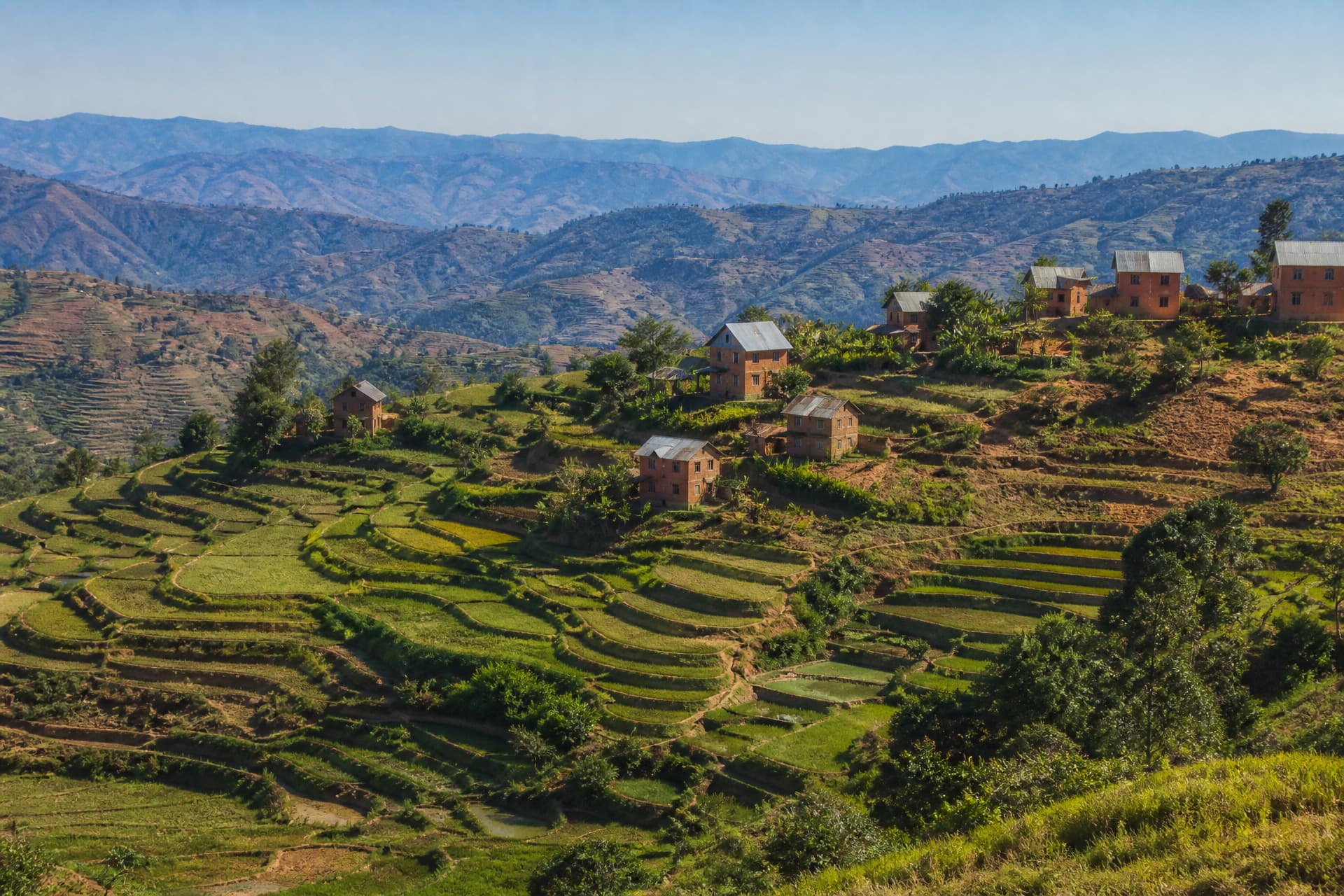 Terraced green hillsides and scattered red-earth houses of a Madagascar highland village photographed in clear dry-season daylight