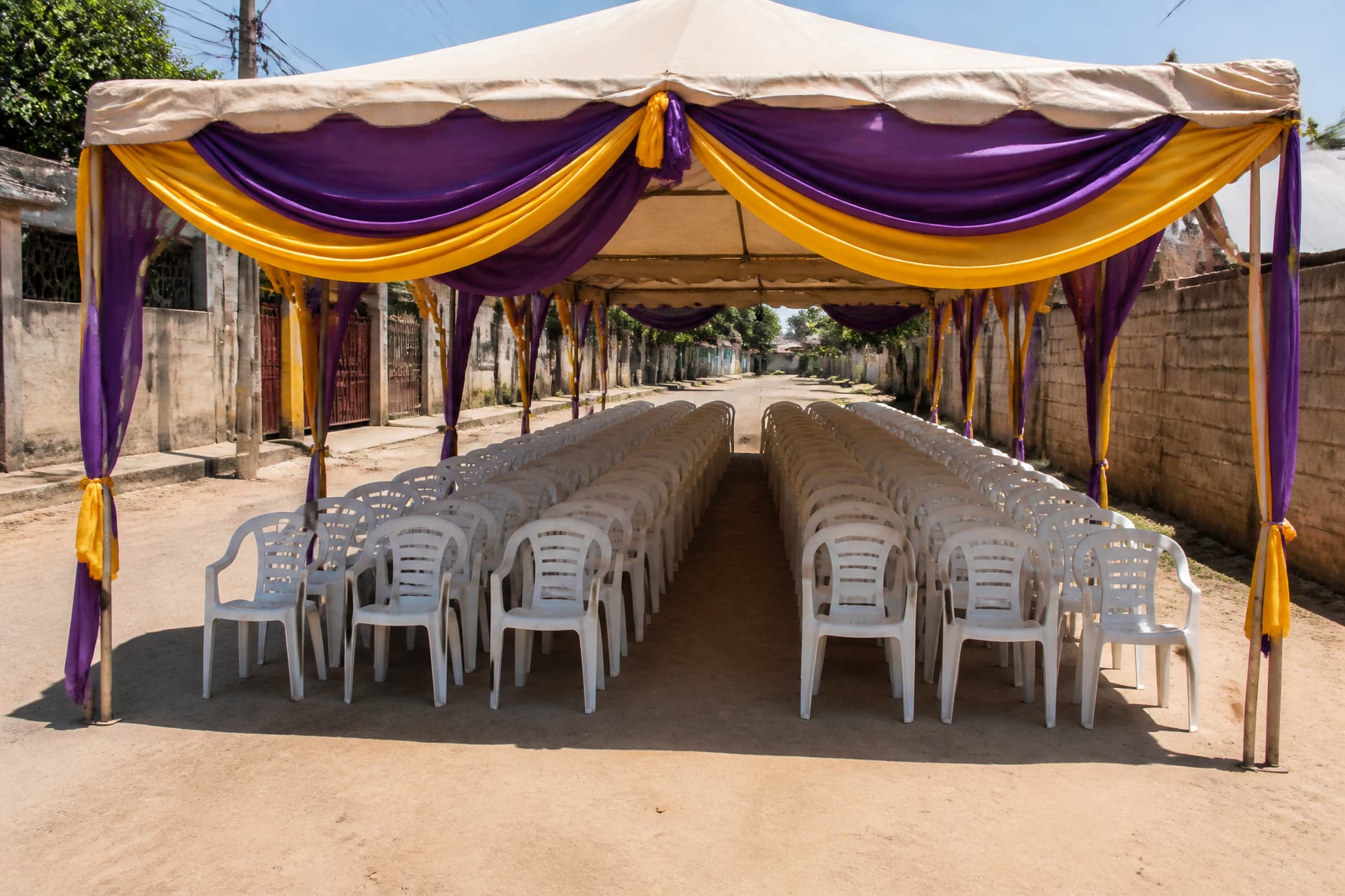 An open-air funeral setting prepared on a dusty street โ a fabric canopy, rows of empty chairs and bright draped cloth, with no people present