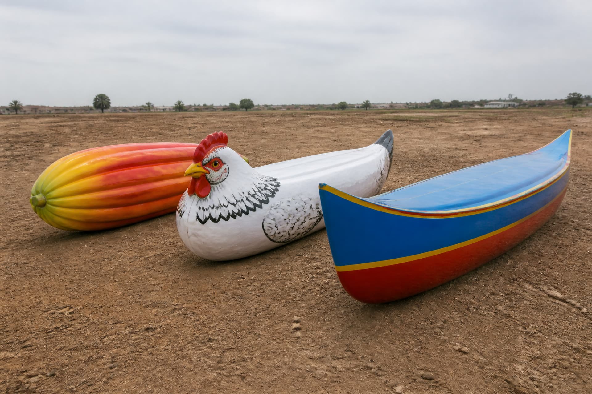 Three finished figurative coffins standing side by side in an open yard โ a cocoa pod, a hen, and a canoe โ painted in bright colors against a plain sky