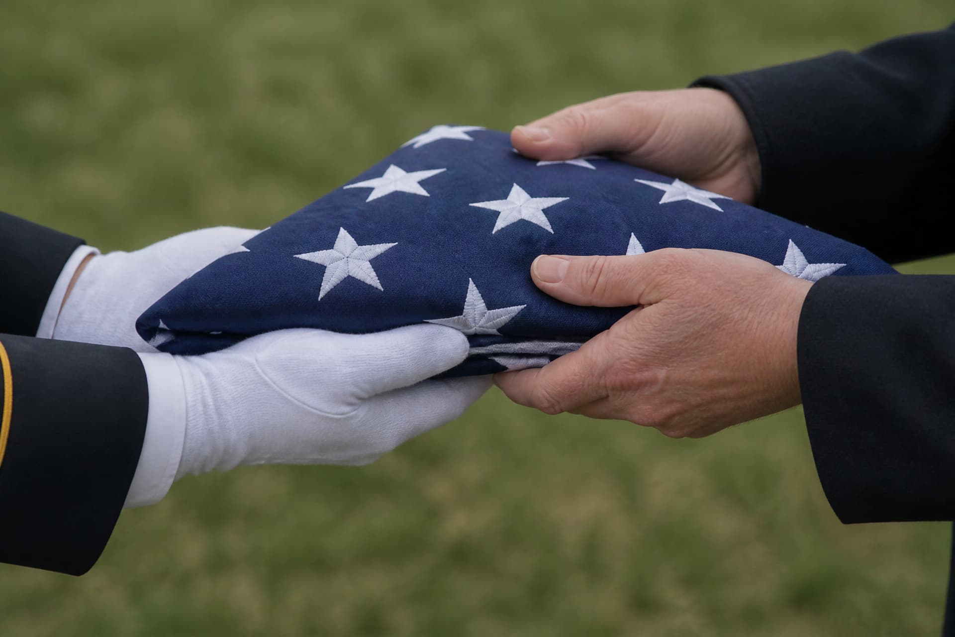 A close view of a folded triangular United States flag passing between two people at a graveside, a pair of white-gloved hands supporting it from below and a pair of bare hands taking hold of it, the straight edge toward the recipient