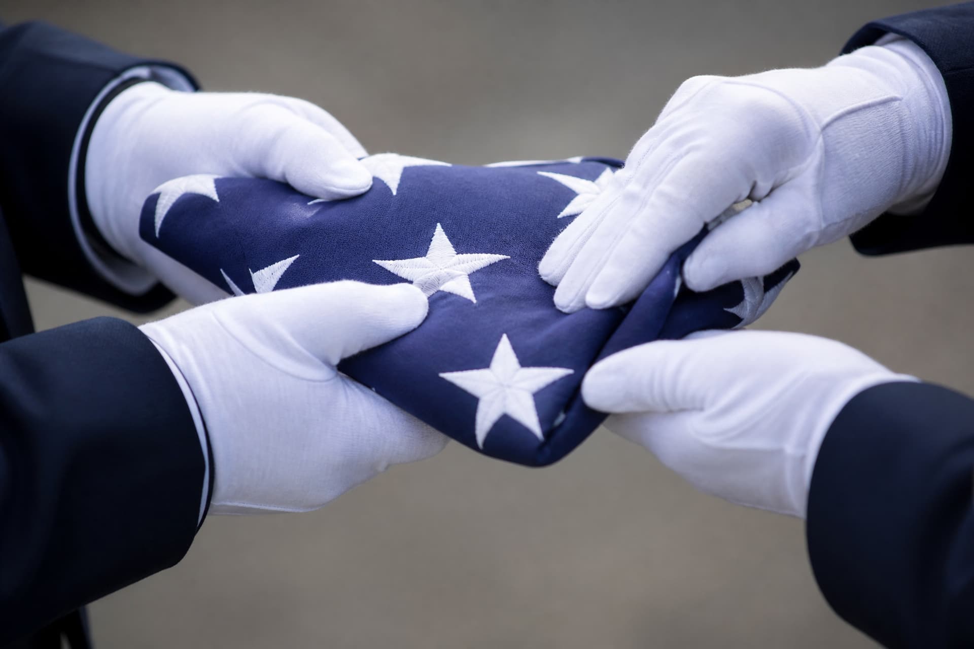 A close view of two pairs of white-gloved hands making the final tuck of a folded triangular United States flag, only the blue field with white stars showing