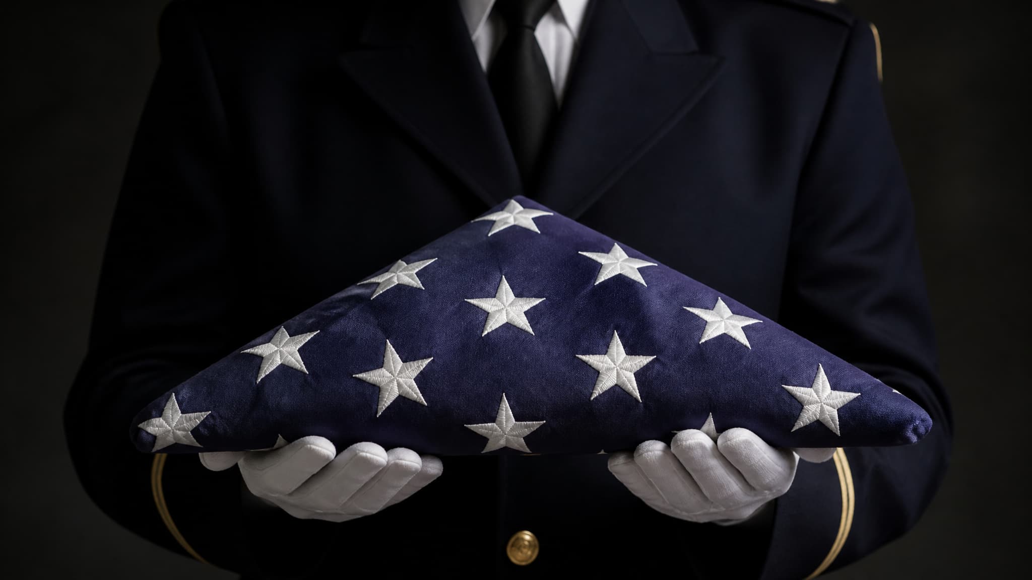A folded triangular American flag held out on two white-gloved hands at chest height by a service member in dark dress uniform, the blue field with white stars uppermost and no stripe showing, face out of frame