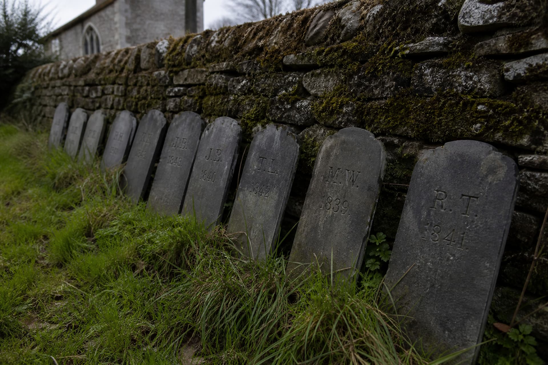 Eleven small weathered slate footstones removed from their graves and stood in a row against a moss-covered dry stone wall in an English churchyard, their carved initials and dates still legible