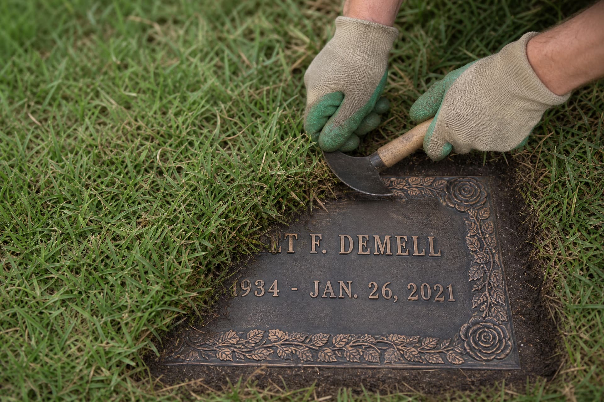 Gloved hands using a half-moon edging tool to cut overgrown turf back from the border of a flat bronze grave marker, one half of the border cleared and sharp and the other still hidden under a lip of creeping grass
