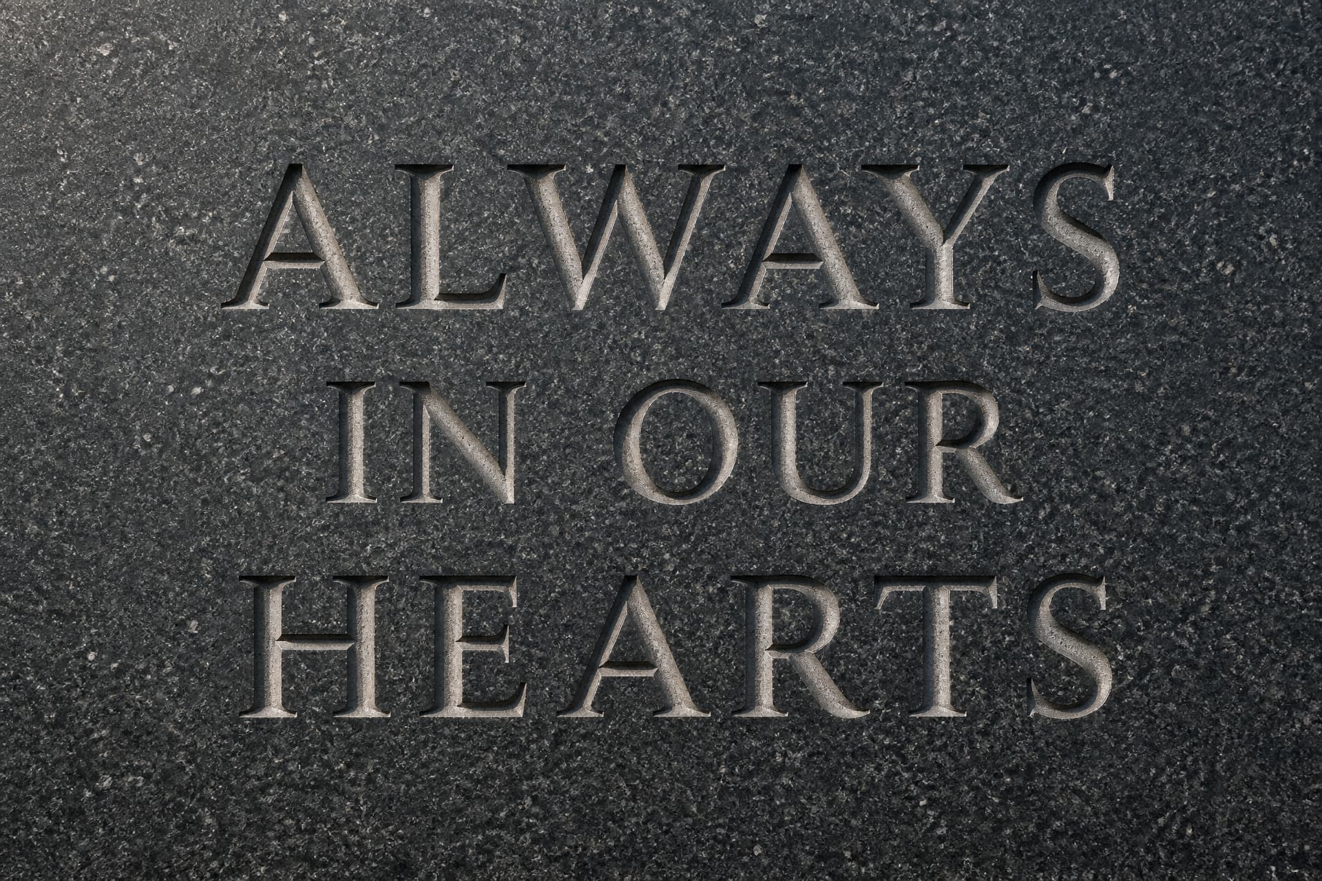 Close-up of a polished dark gray granite memorial face with ALWAYS IN OUR HEARTS sandblasted into it in three lines of recessed serif capitals