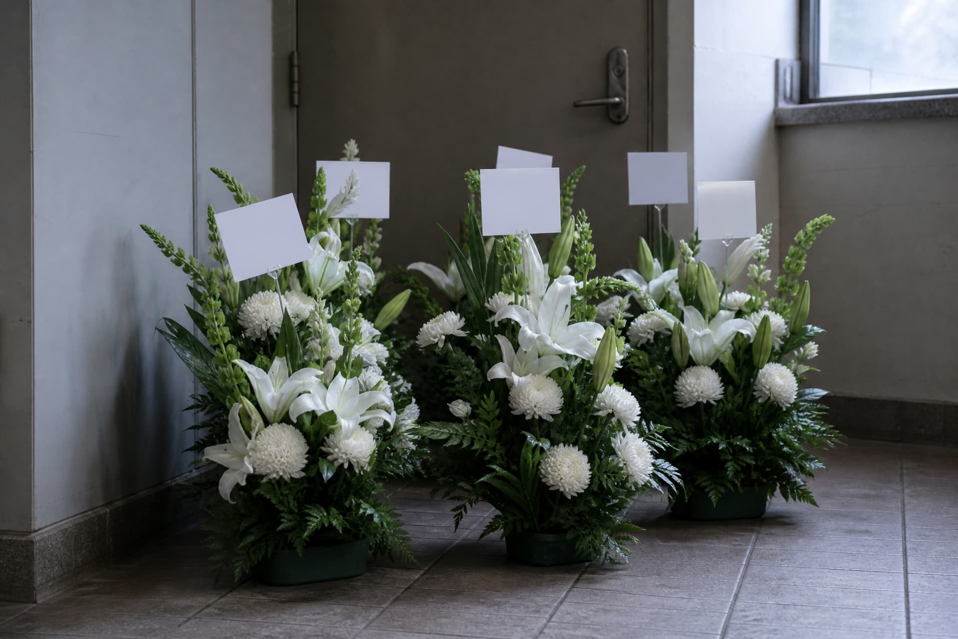 Several funeral flower arrangements of white lilies and chrysanthemums standing together on a tiled floor just inside a service entrance, a small blank card clipped to a stick in each one